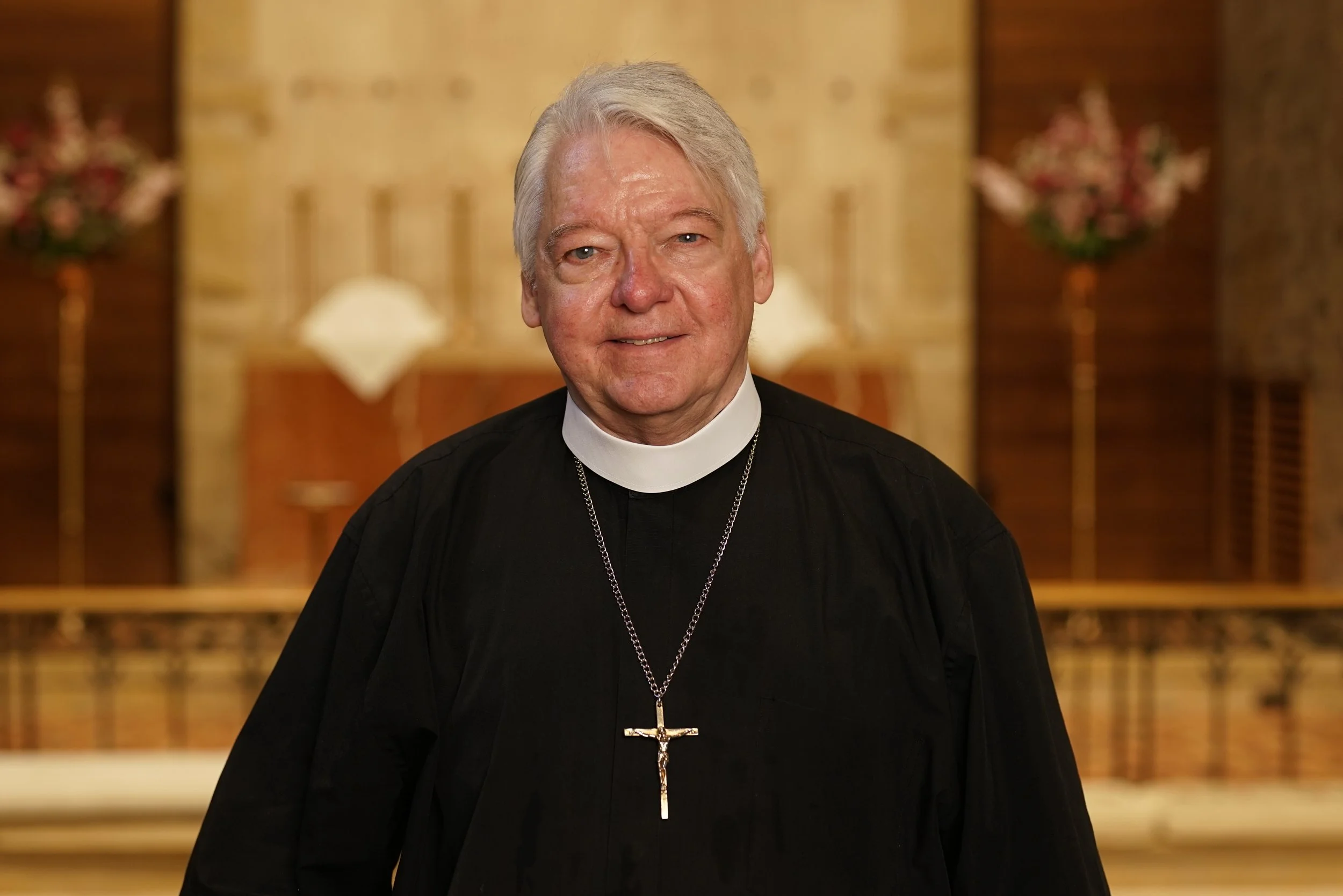 An elderly man dressed as a pastor, wearing a black robe and a silver cross necklace, standing inside the church sanctuary.
