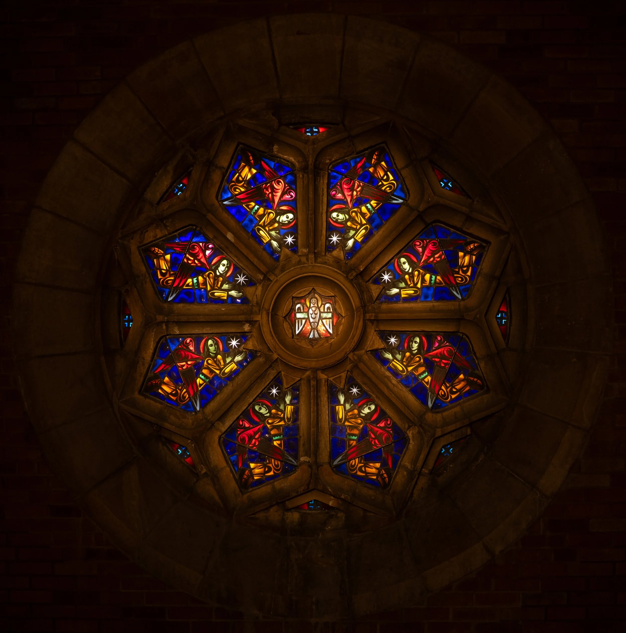 Colorful stained glass rose window with a central figure of the Holy Spirit as a dove, and surrounding angels holding eight-pointed stars (blessings)