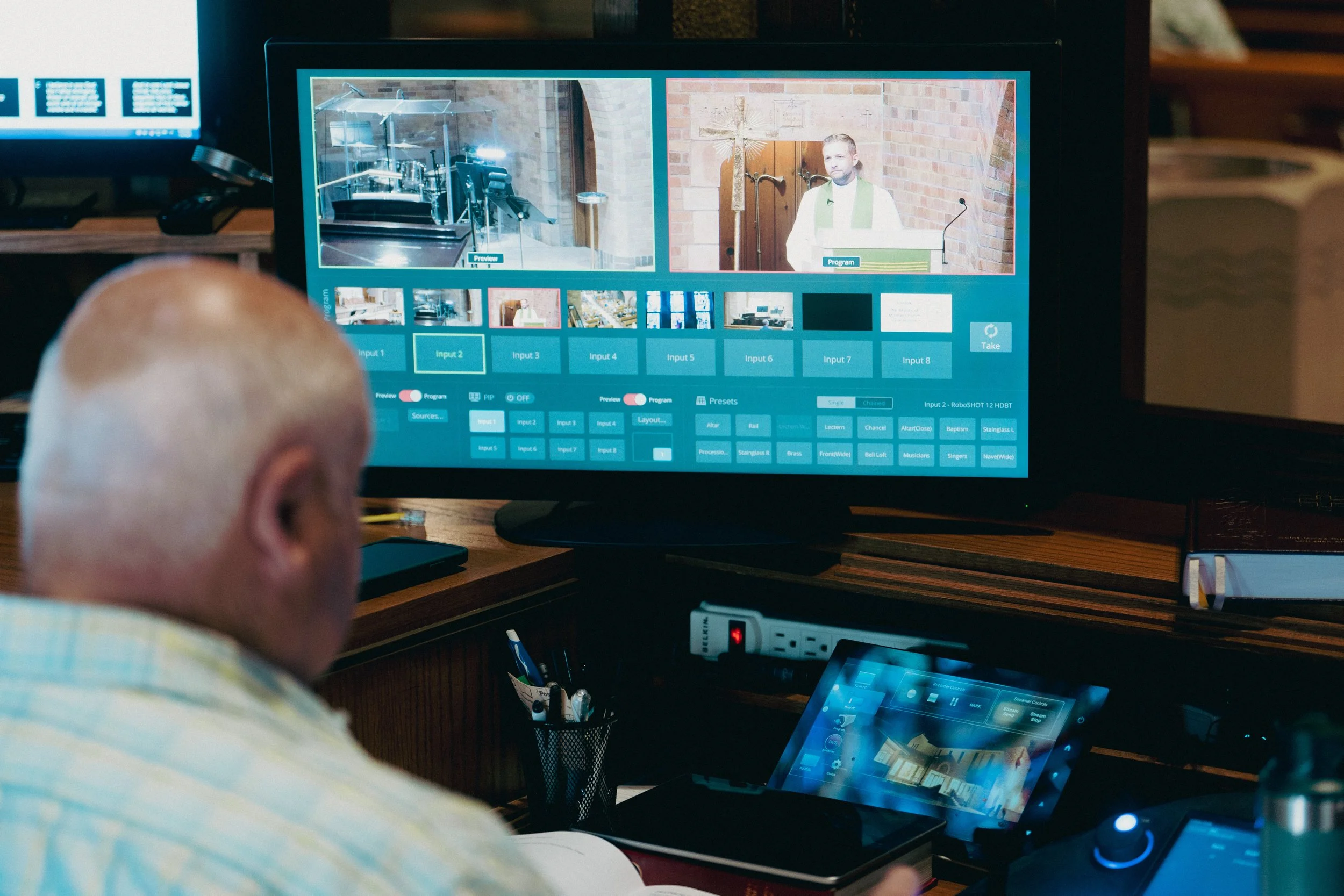 A man with a shaved head is operating a video switcher with multiple input feeds, displayed on a large screen in front of him. The screen shows various video inputs, including a religious service with a pastor and a church interior.