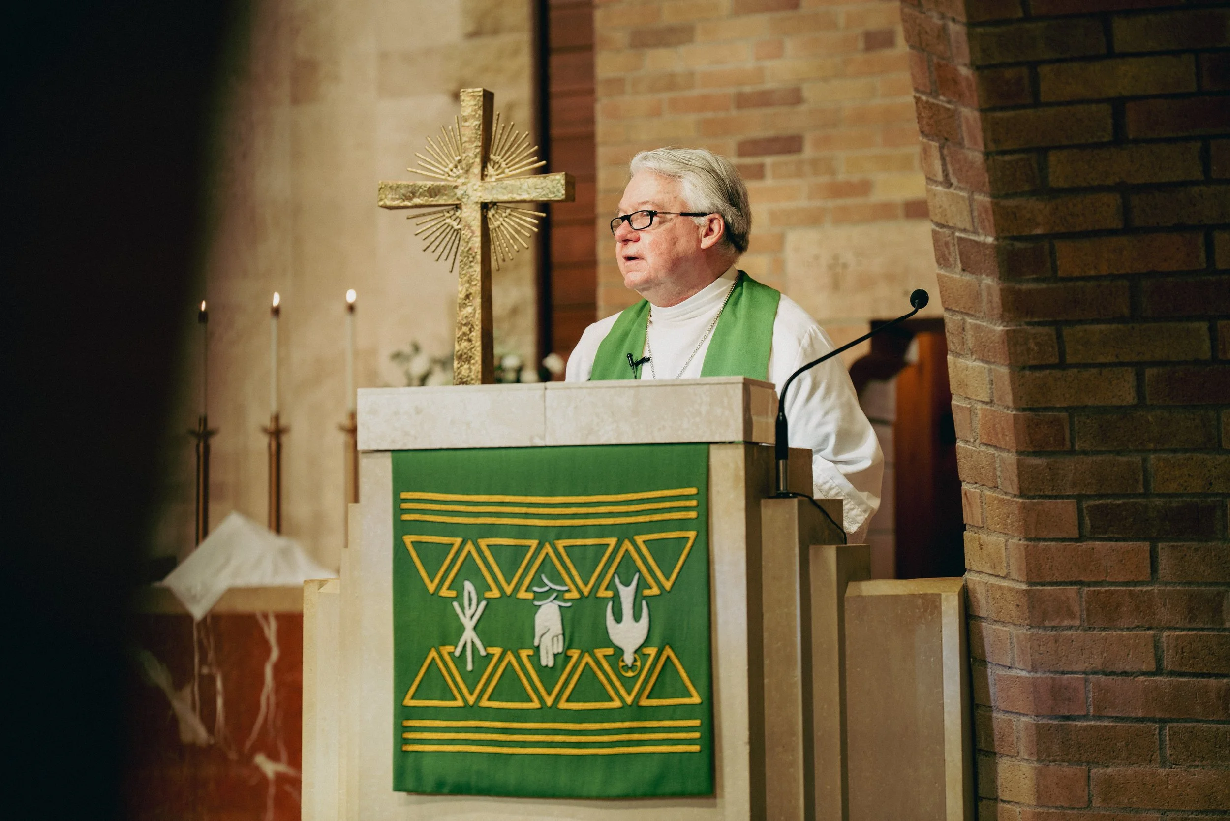 A pastor standing at a pulpit in church, giving his sermon. The pulpit is decorated with a green cloth featuring Christian symbols. Behind him are brick walls, lit candles, and a gold cross.