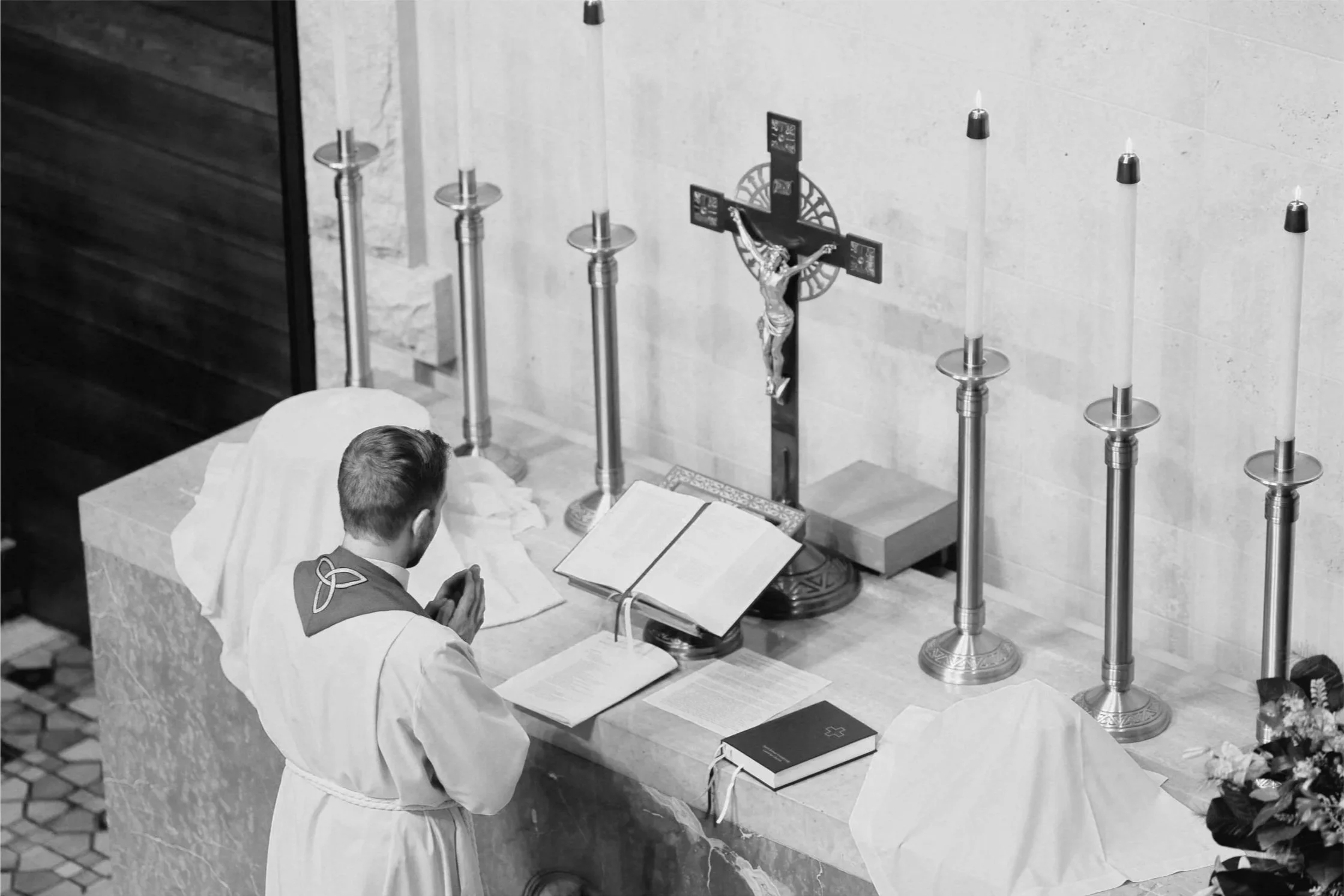A pastor in prayer at an altar with an open Bible, candles, a crucifix, and a cloth.