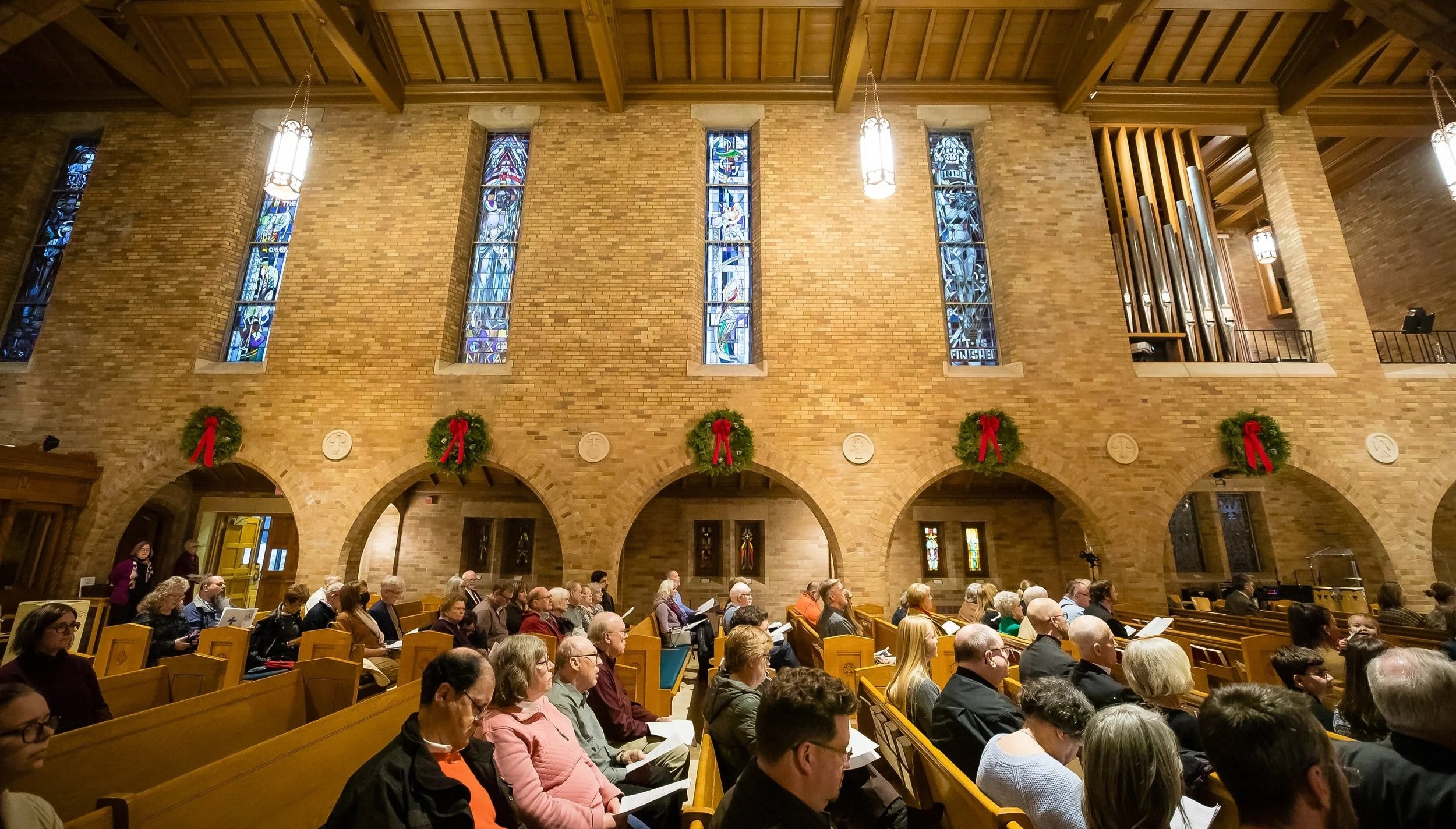 People seated in church during a service, with stained glass windows on a brick wall and Christmas wreaths with red ribbons decorating the space.