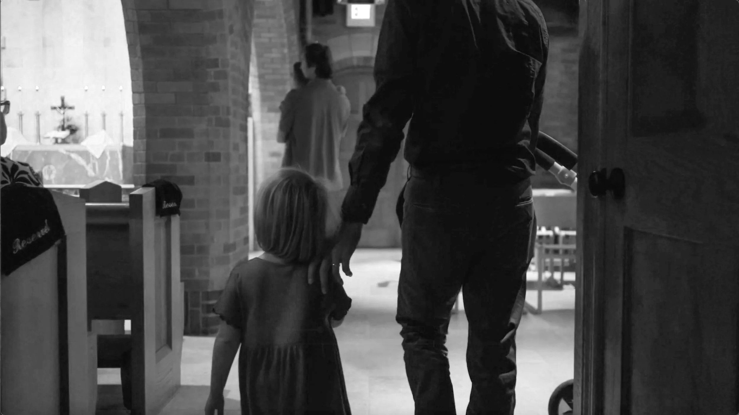 A father is gently holding the hand of a young girl while walking down the side aisles of the pews.