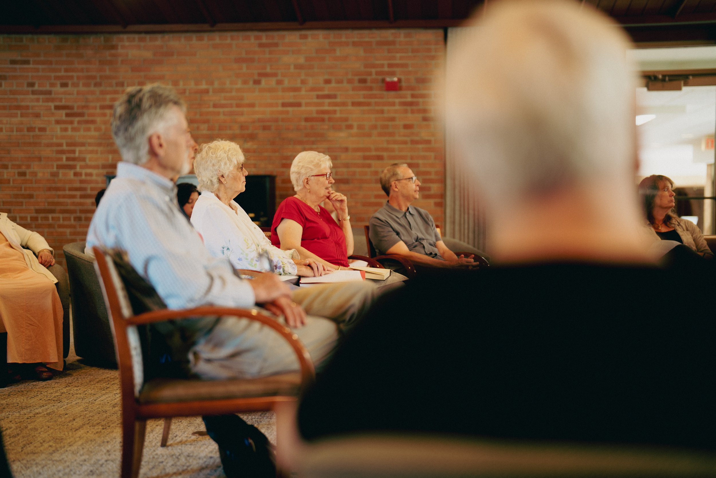 A group of older adults seated in chairs, attentively listening to a speaker or presentation in an indoor setting with a brick wall background.