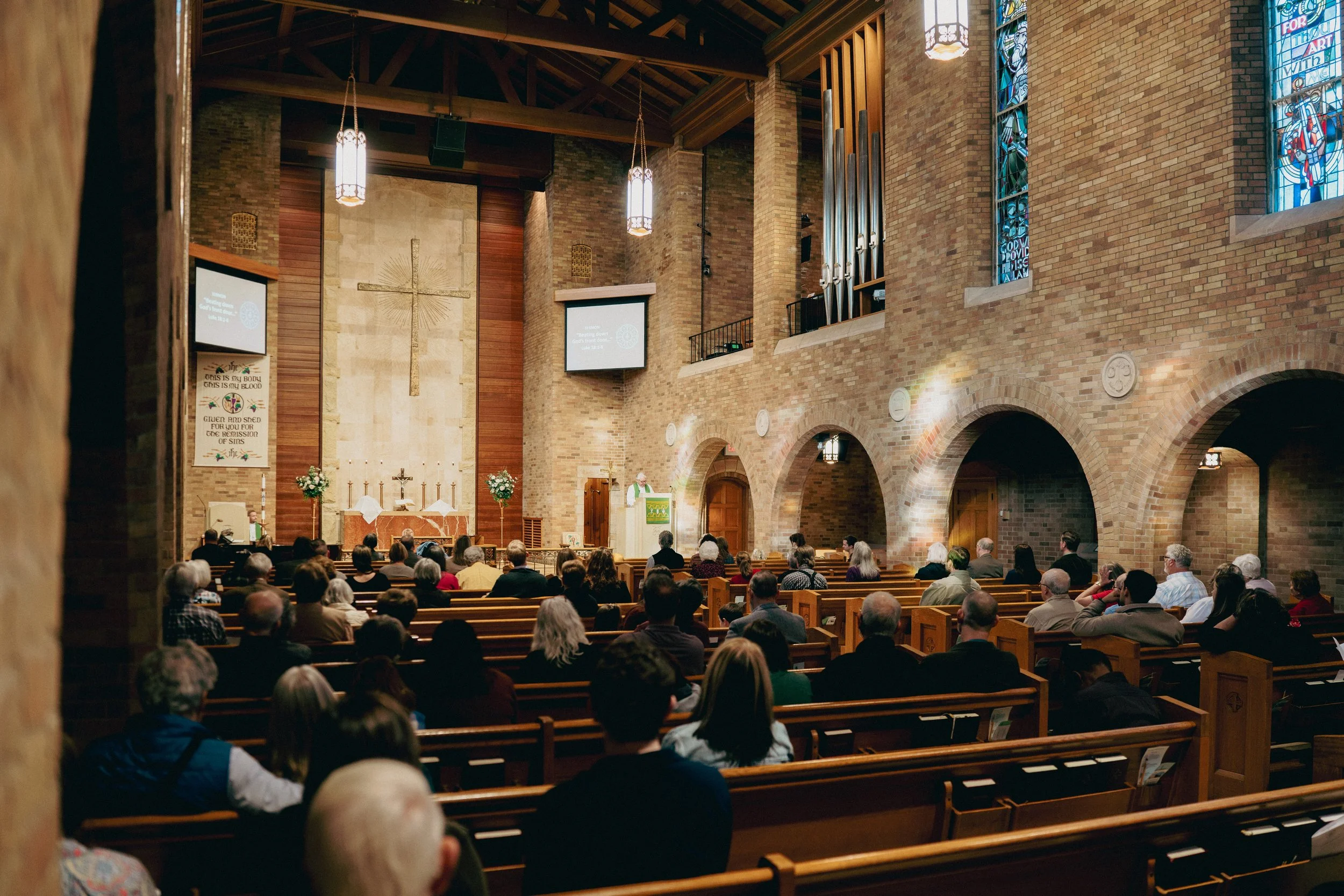 Inside the church sanctuary filled with people seated on wooden pews, with stained glass windows on brick walls, a cross above the altar, and a pastor speaking at a pulpit.