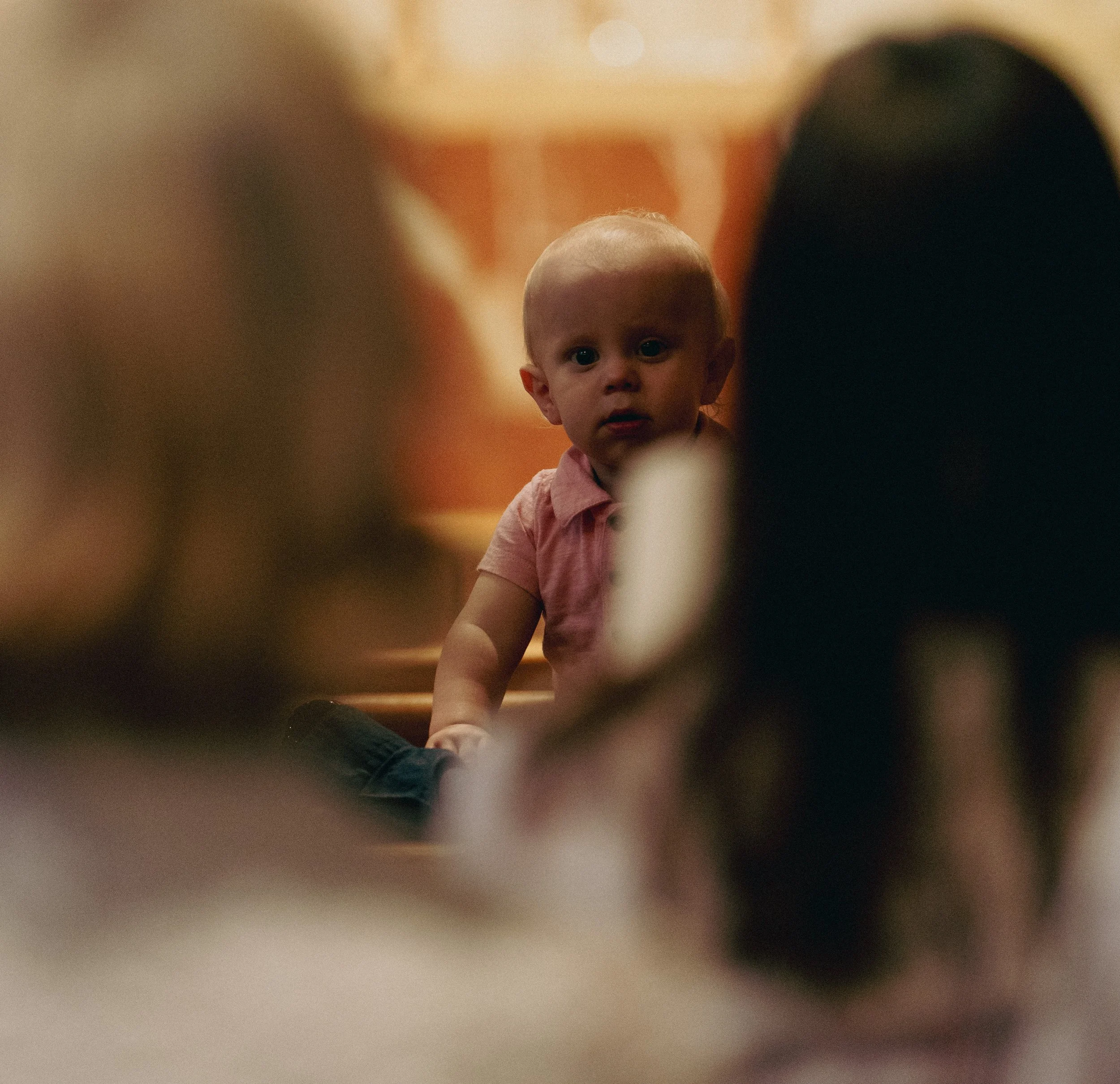 A baby looking over their parent's shoulder directly at the camera with a curious expression, framed by blurred figures in the pews.