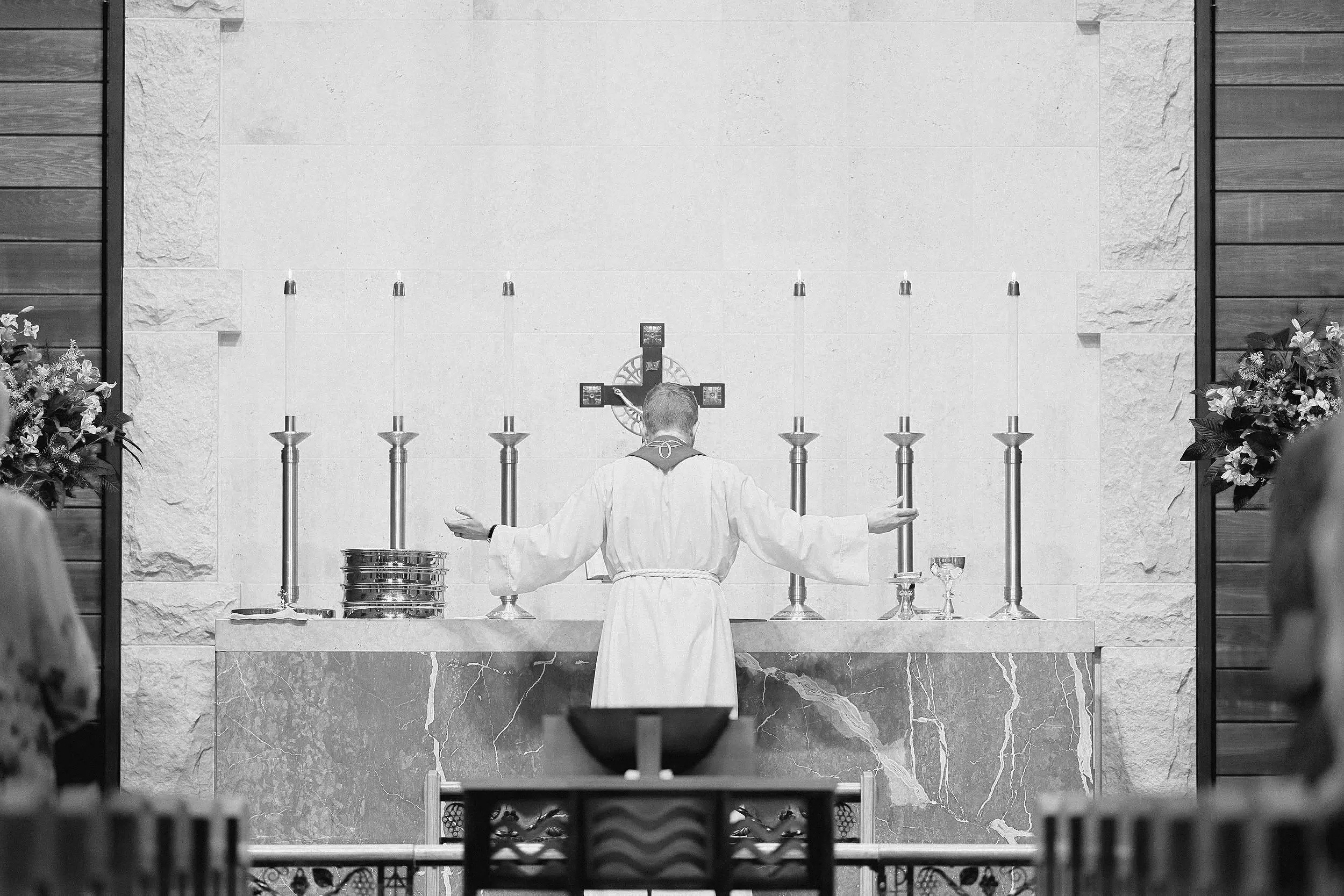 A black and white photo of a pastor standing at the altar with arms outstretched during a church service, facing a cross with candles on either side.