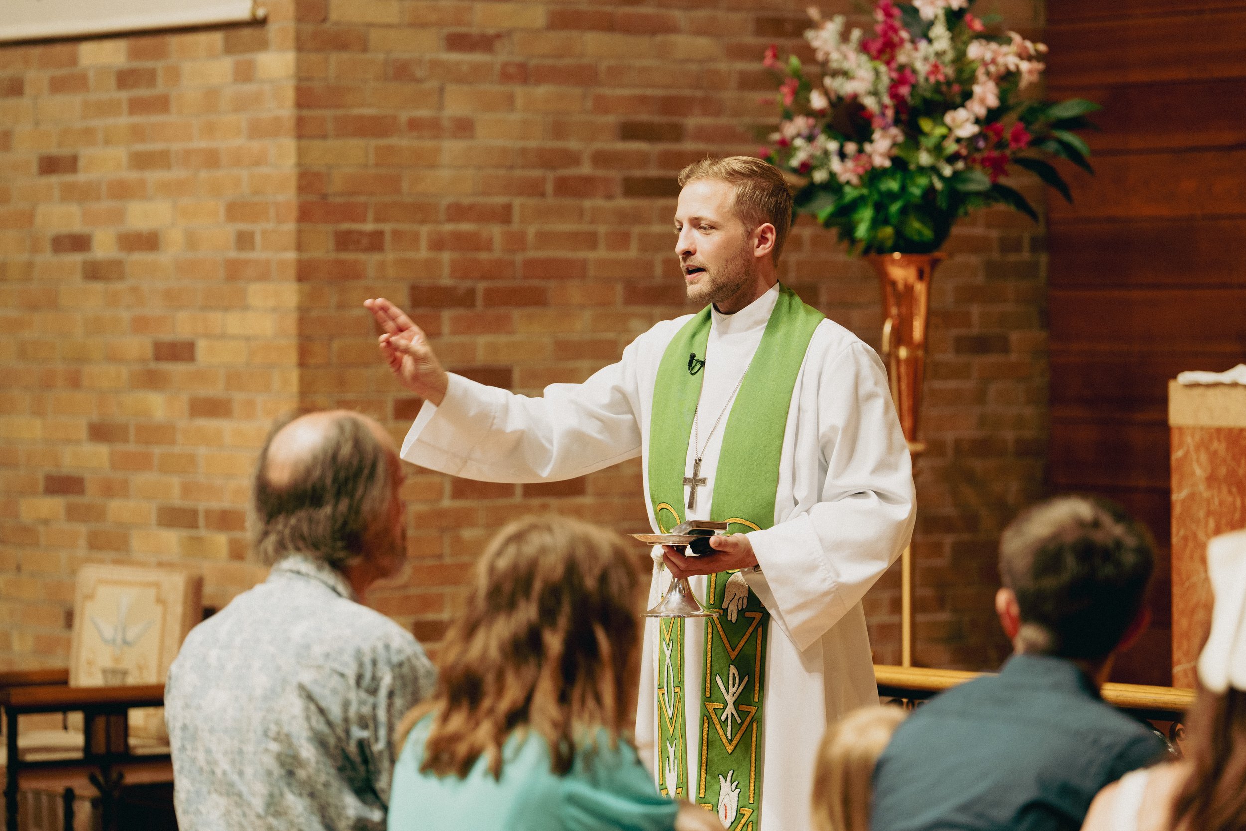 A pastor in white robes with a green stole and cross necklace performs a blessing in front of congregation members at the communion rail, inside a church with brick walls and a large flower arrangement in the background.