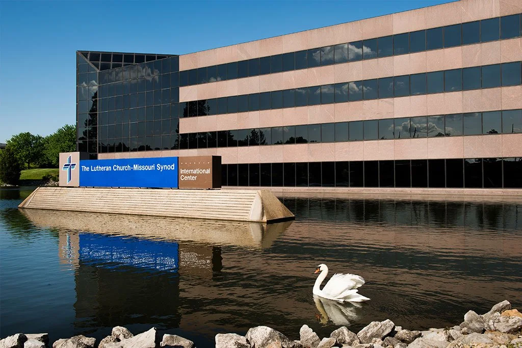 Modern office building with reflective glass windows beside a pond, with a swan swimming in the water and rocks along the shoreline. A sign on the building reads 'The Lutheran Church-Missouri Synod International Center'.