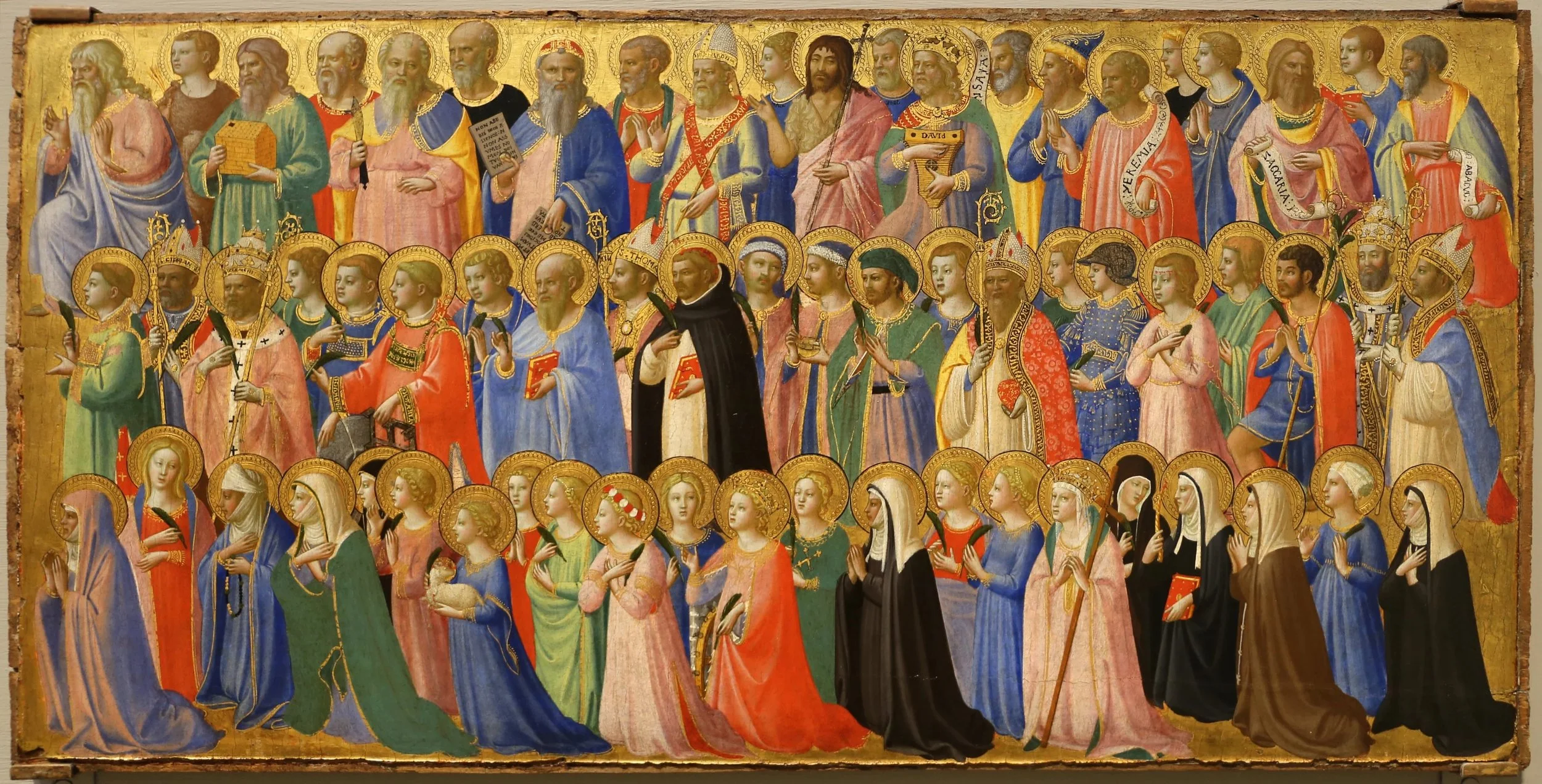 A detailed medieval religious painting featuring numerous saints, apostles, and religious figures, many with halos, depicted in vibrant robes, some holding scrolls, crosses, or other religious symbols, set against a gold leaf background.