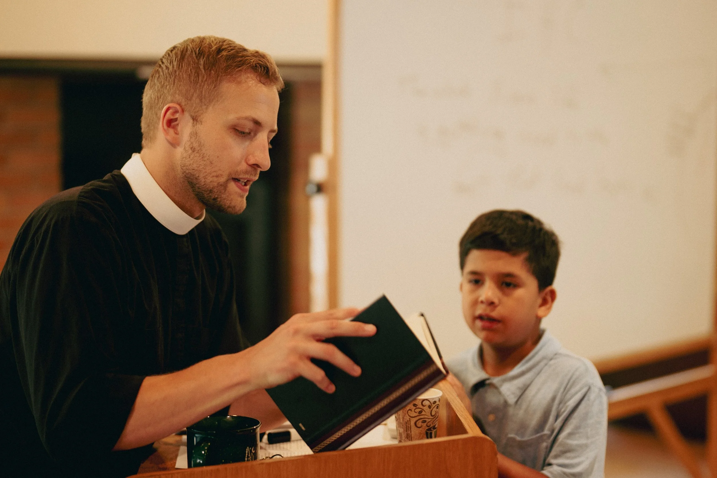 A man dressed as a pastor in a black shirt with a white collar sitting at a table with a young boy, holding a book and engaging in conversation.