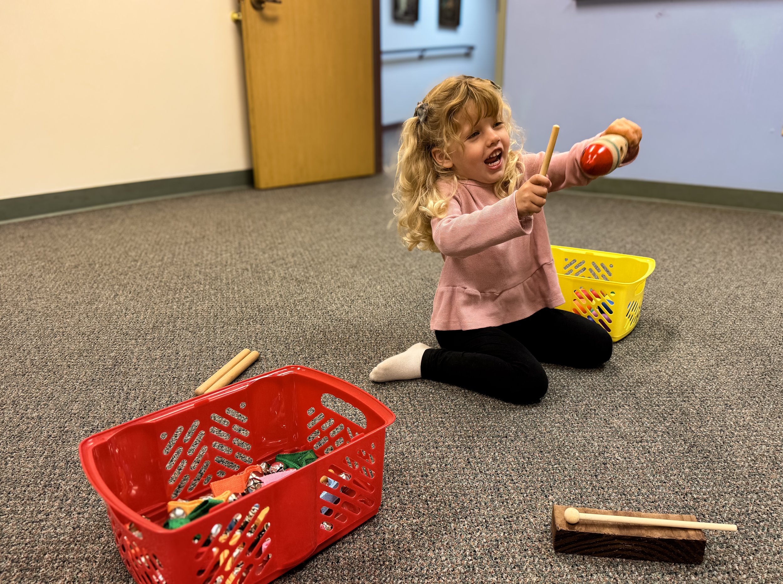 A young girl with blonde curly hair, wearing a pink sweatshirt and black pants, is sitting on the floor of a classroom holding a wooden mallet and striking a toy drum. She appears happy and engaged. There are two baskets, one red and one yellow, filled with various toys and percussion instruments nearby. The classroom has beige carpet, a closed yellow door, and a wall with light blue and white colors.