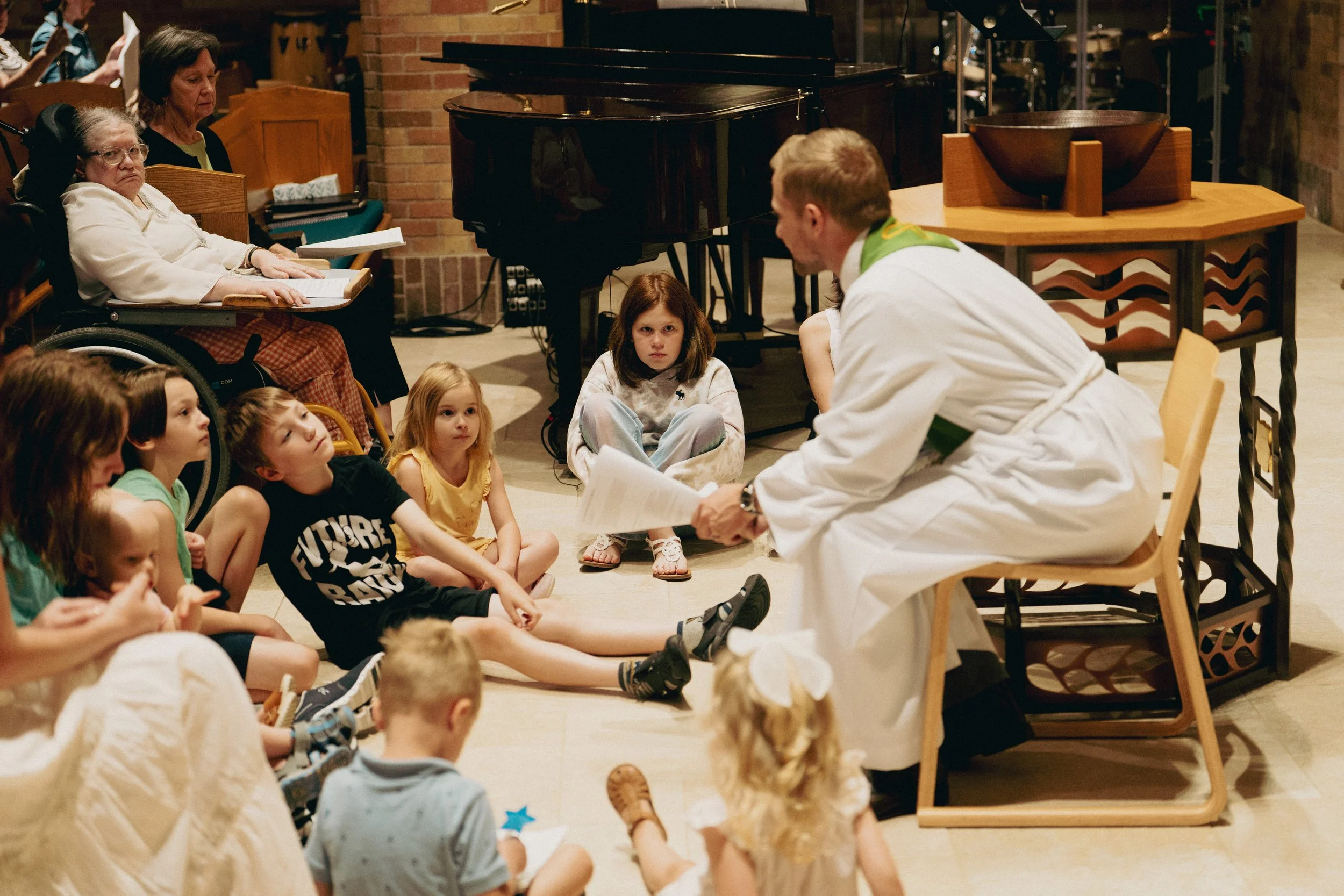 A man dressed as a pastor talking to a group of children sitting on the floor, with an elderly woman in a wheelchair and other adults in the background.