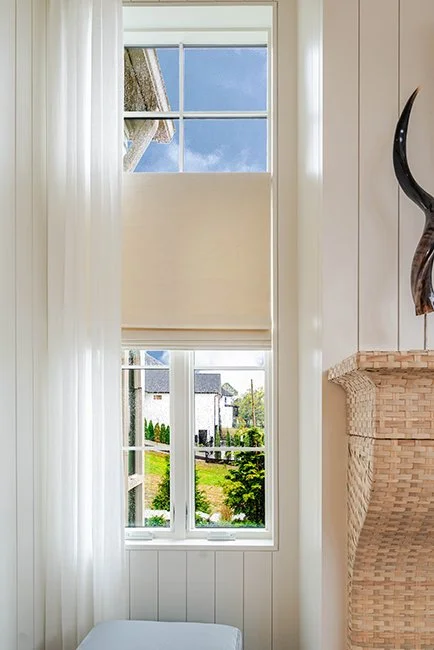 Interior view of a house showing a large, double-pane window with white sheers and shade, overlooking a sunny backyard with green trees and neighboring houses.