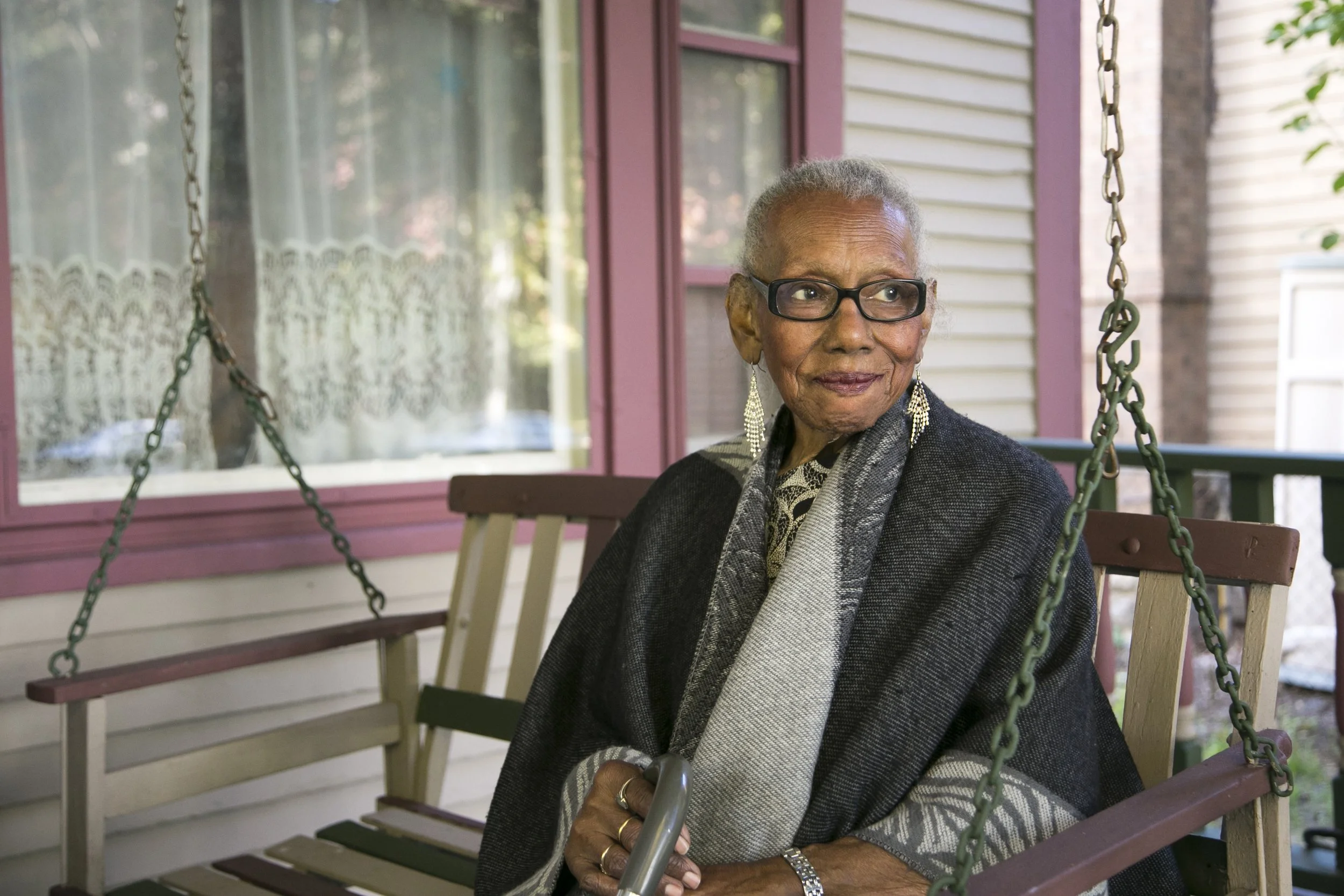 A woman with dark skin tone wearing a gray coat smiles as she sits on a wooden swing on the front porch of her home.