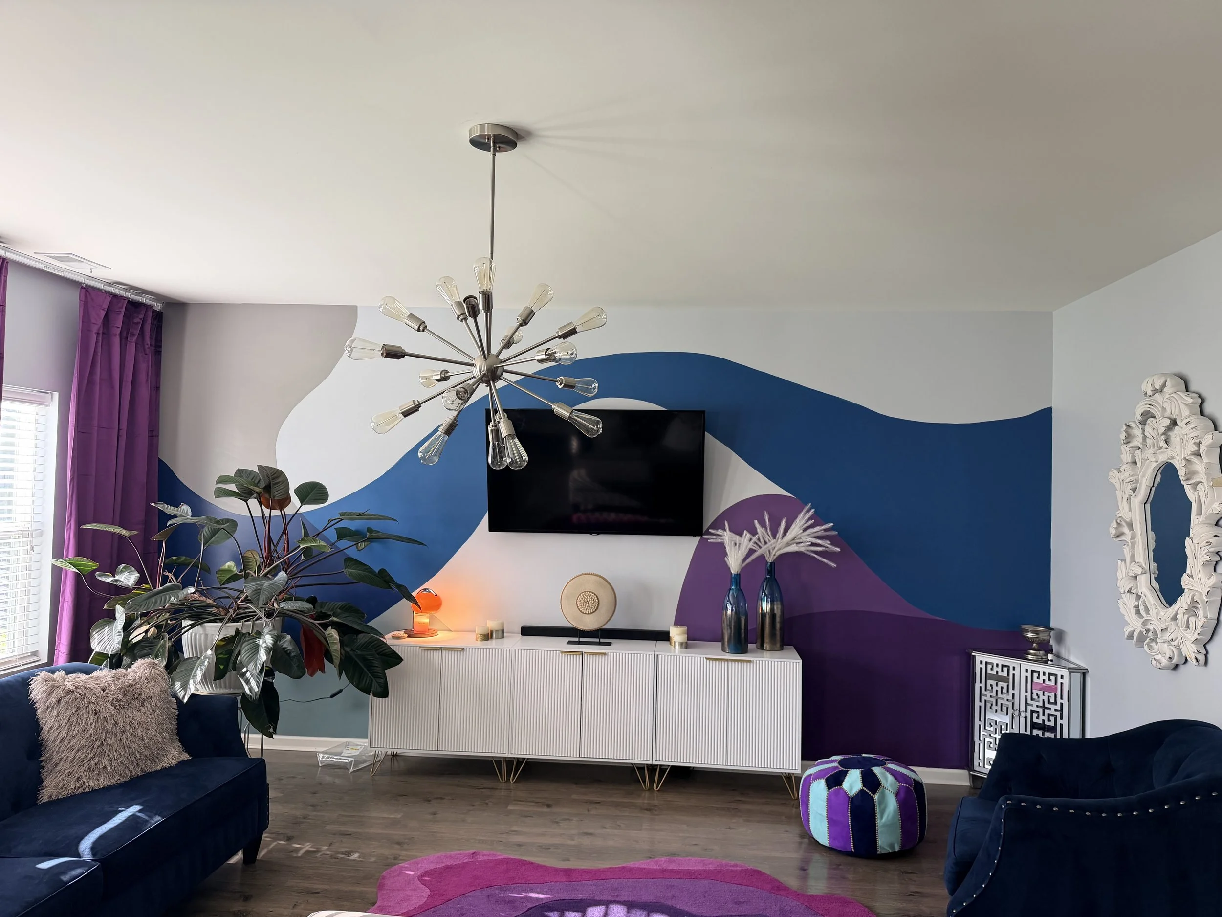 This living room wall didn't need more furniture. It needed movement.
A sweeping abstract mural in navy, violet, and soft white transforms this feature wall into the anchor of the entire space — flowing around the television, pulling the eye across t