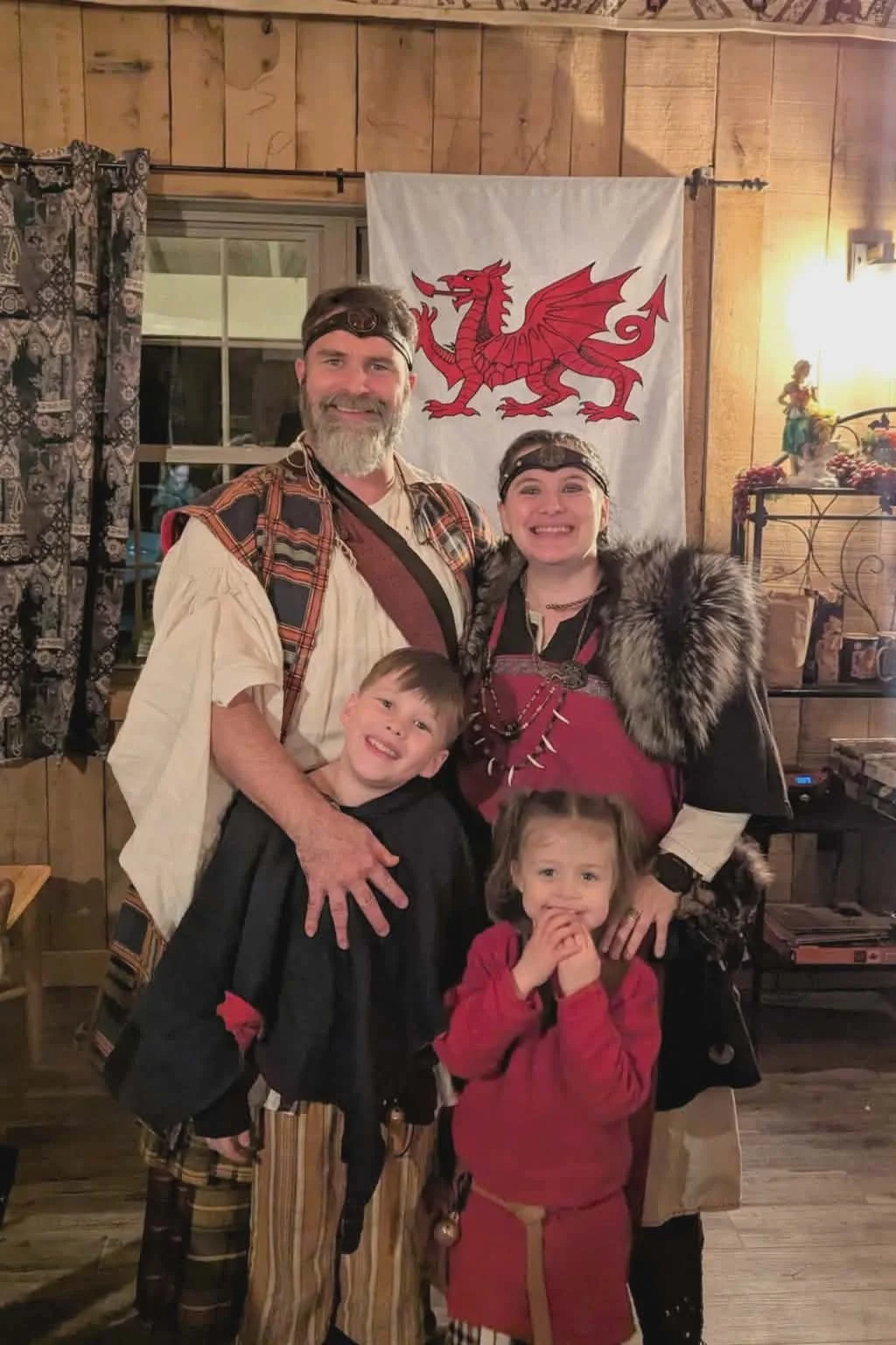 Family dressed in medieval costumes, smiling, standing in front of a Welsh flag with a red dragon, in a warmly lit room with wood-paneled walls and decorative elements. Yule Festival with Viking kids. ReddWolf
