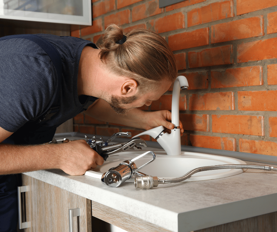 Plumber installing a kitchen faucet with tools on a countertop in front of a brick wall.