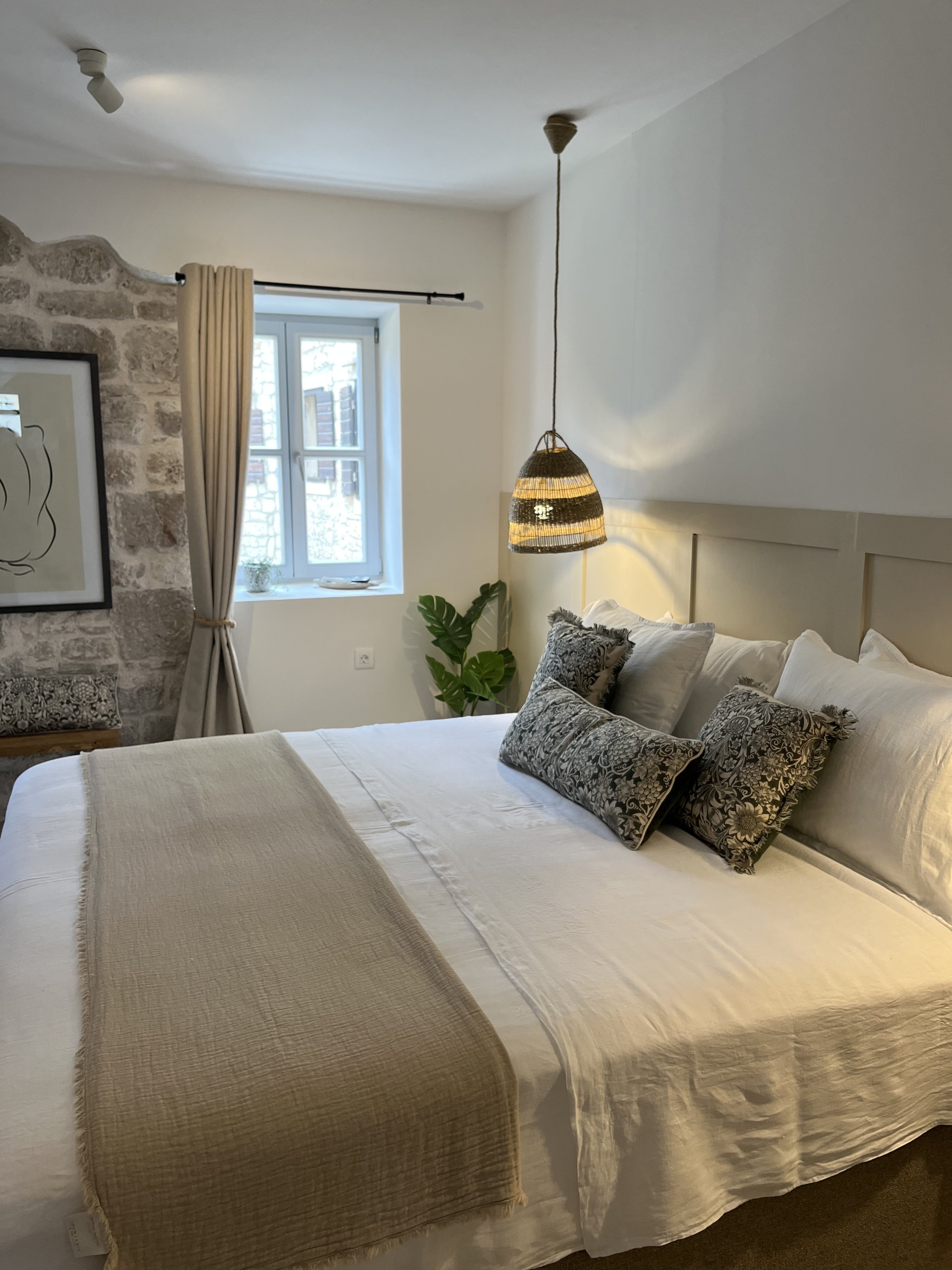 A cozy bedroom with a white bed, decorative pillows, a beige bed runner, a hanging pendant light, a window with curtain, a plant, and a framed drawing on the wall.