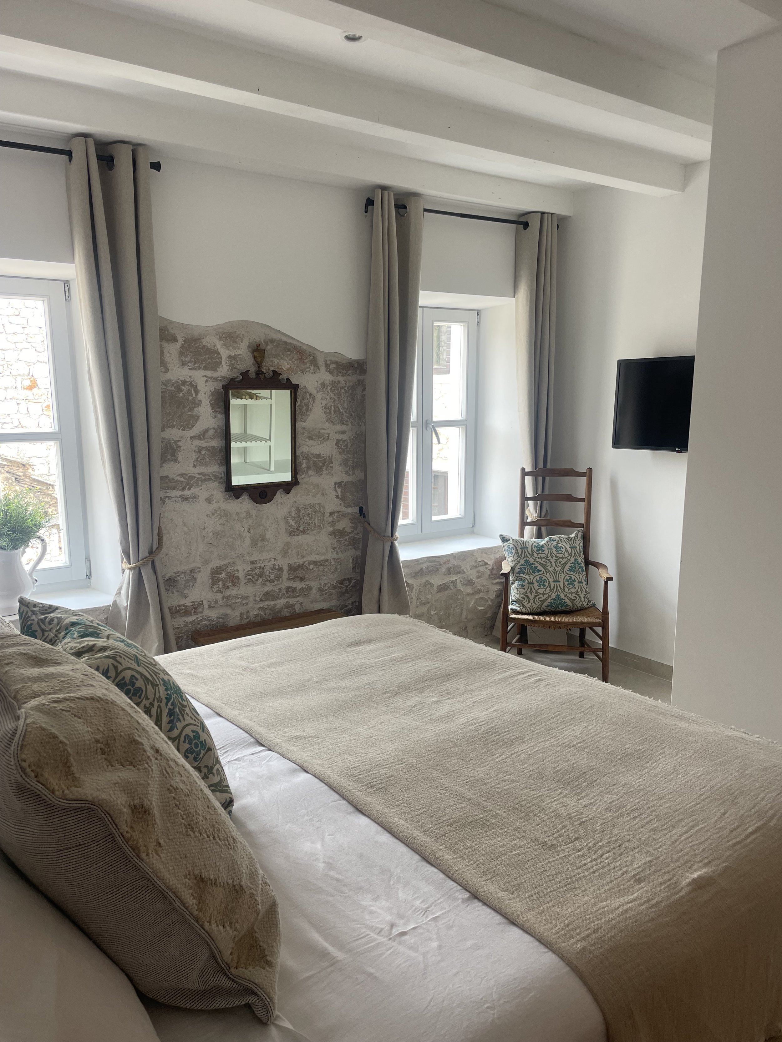 A cozy bedroom with two windows, beige curtains, a bed with decorative pillows, a small mirror on a stone wall, a wooden chair with a pillow, and a mounted flat-screen TV.