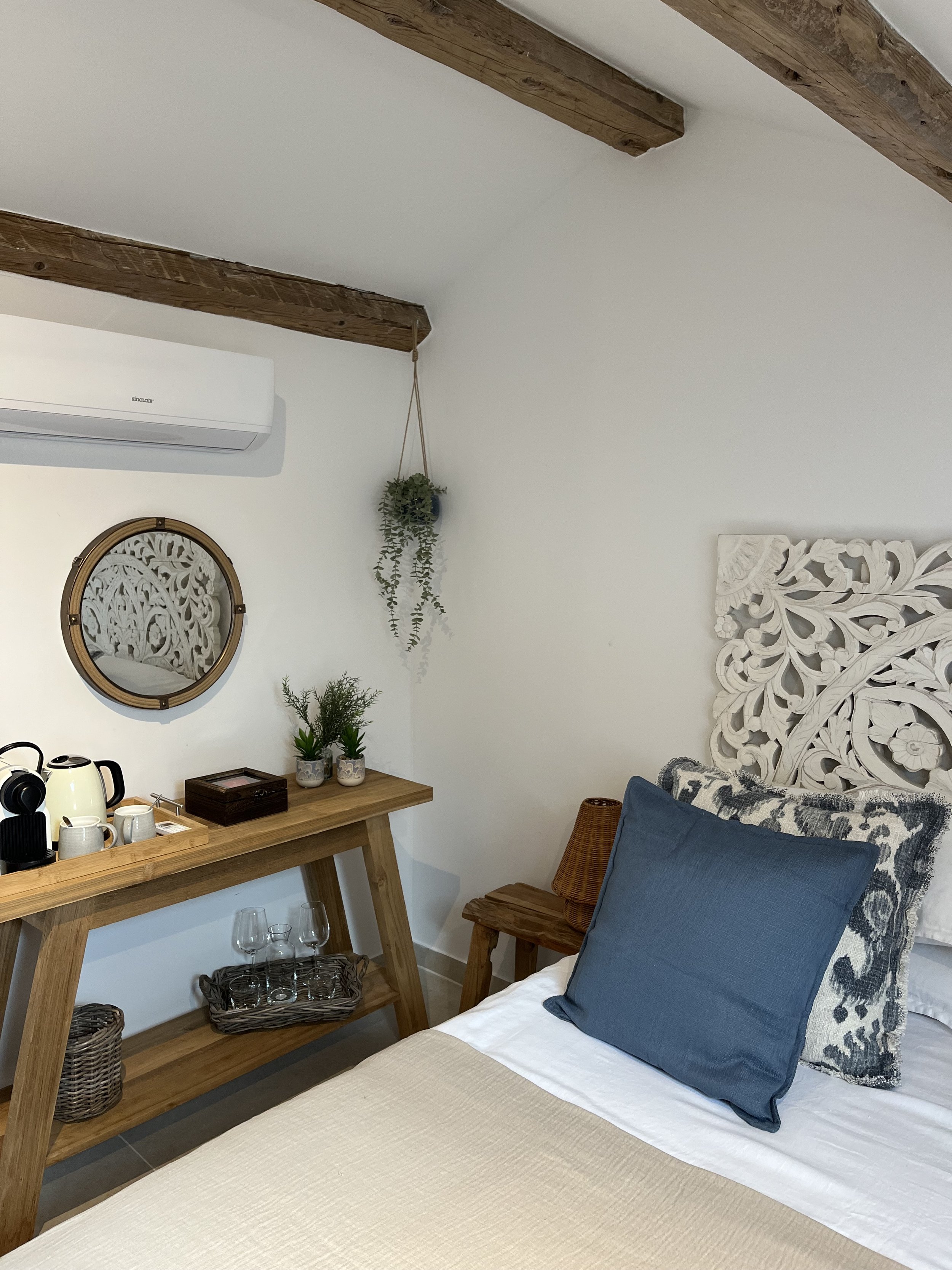 Cozy bedroom corner with a bed featuring a white carved headboard, blue and patterned cushions, a wooden side table, and a wooden desk with decorative plants, a mirror, and beverage cups, under wooden ceiling beams.