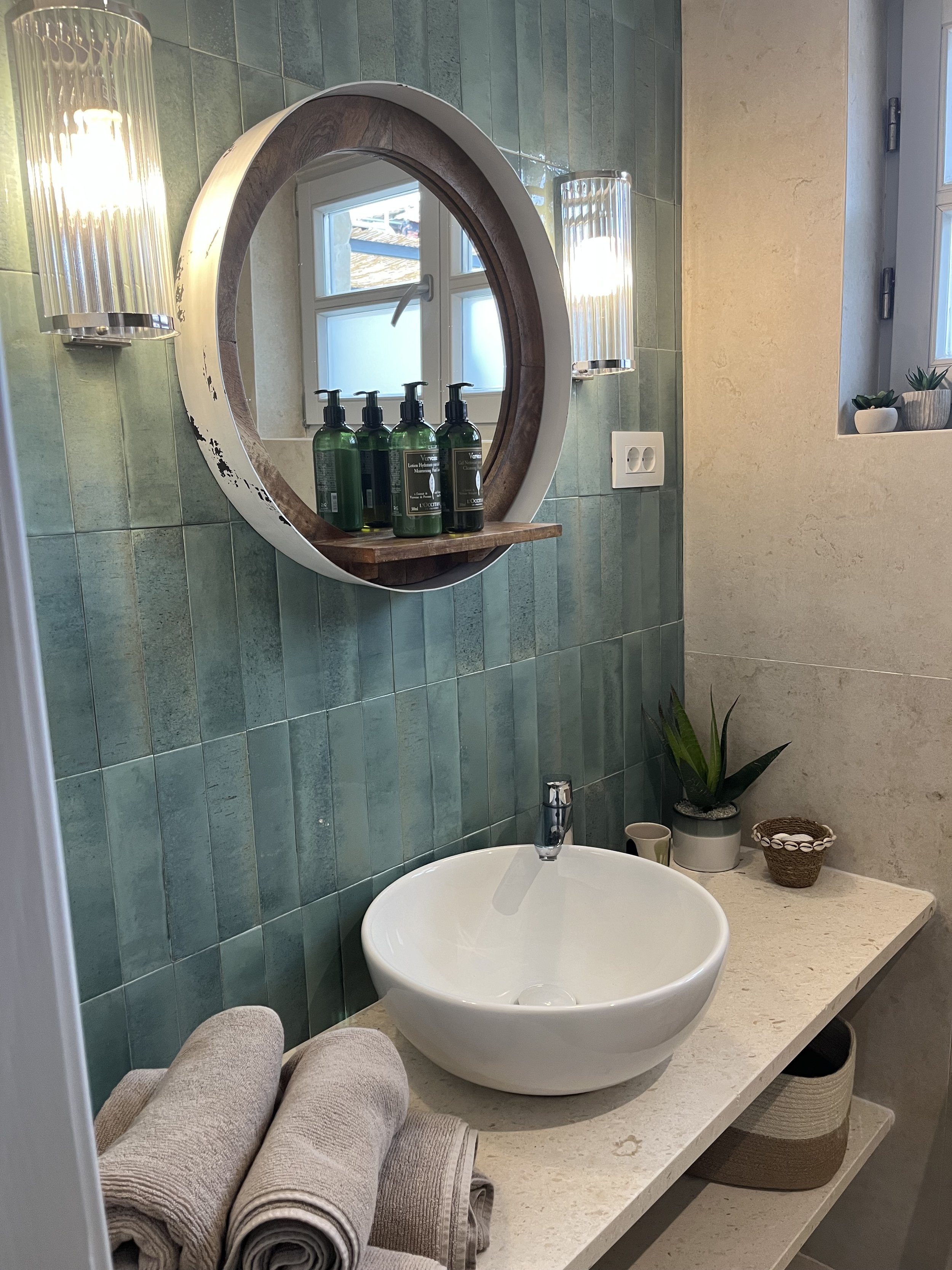 Bathroom with green tiled wall, round mirror with wood and white frame, wall-mounted light fixtures, three green soap dispensers on shelf, white vessel sink, beige towels, small potted plant, candles, and natural decorative items.