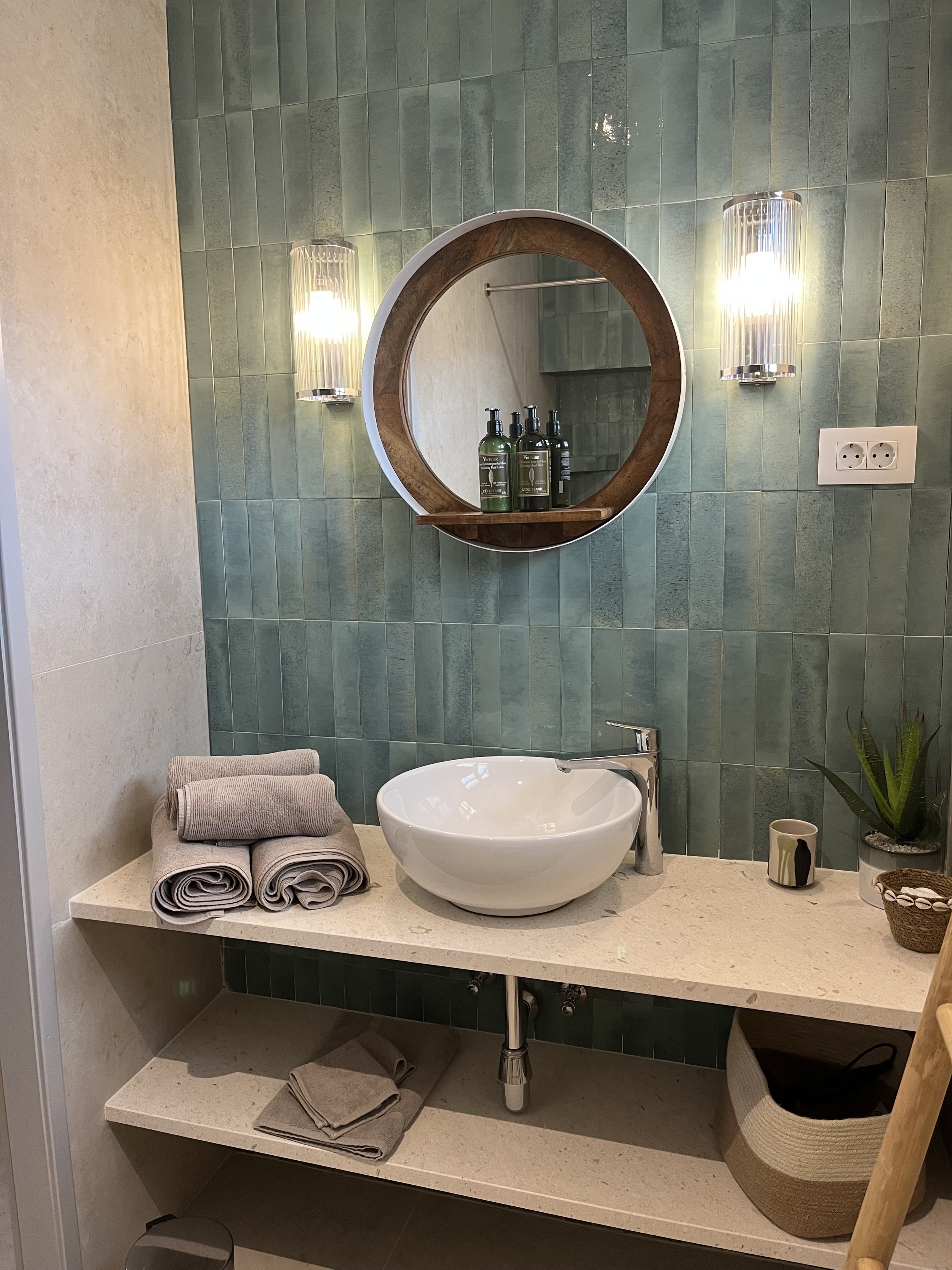 Modern bathroom with a circular mirror, wall-mounted lights, green tiled wall, white vessel sink, beige countertop, and neatly rolled towels.