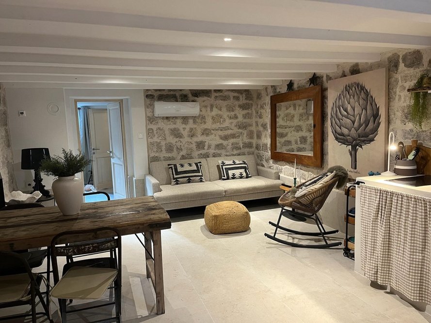 Living room with a stone accent wall, beige sofa with black and white pillows, wooden framed mirror, modern art painting of a flower, rocking chair, small ottoman, large potted plant on a rustic wooden table, and a dining area with chairs.