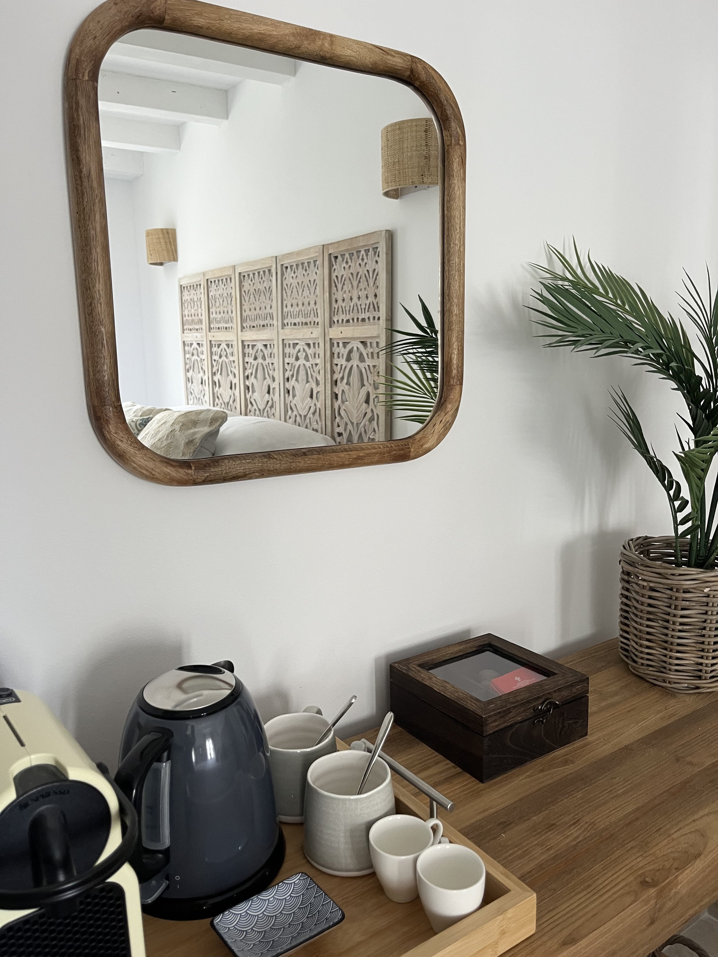 A wooden-framed square mirror reflecting a carved wooden headboard, a wall-mounted wicker lamp, and white walls. On the wooden surface below, there are white ceramic cups, a black kettle, a cream-colored coffee maker, and a potted palm plant.