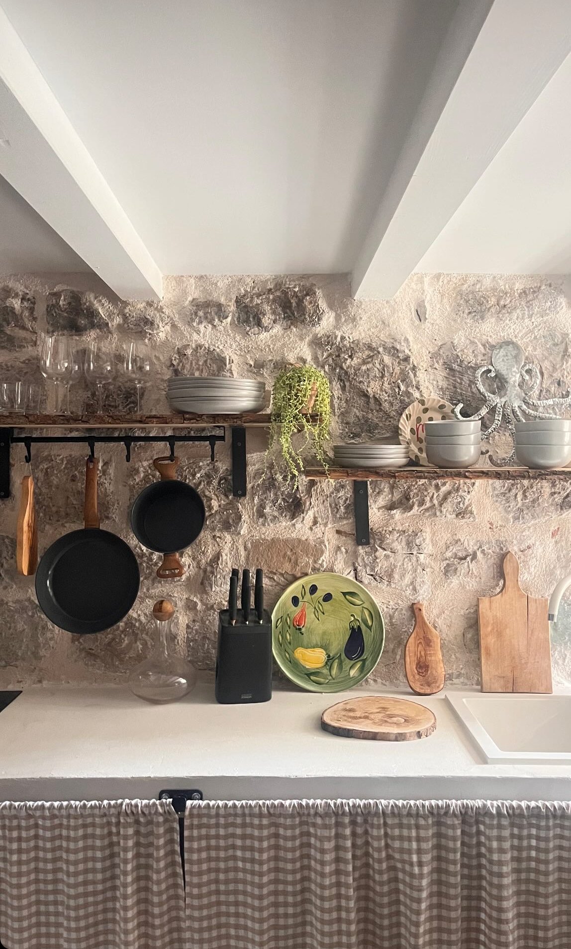 Kitchen with a stone wall backsplash, white countertop, and open wooden shelves holding dishes and glassware. Hanging pots and a knife block are on the counter, with a cutting board and a decorative plate featuring fruit and vegetables. A checkered c