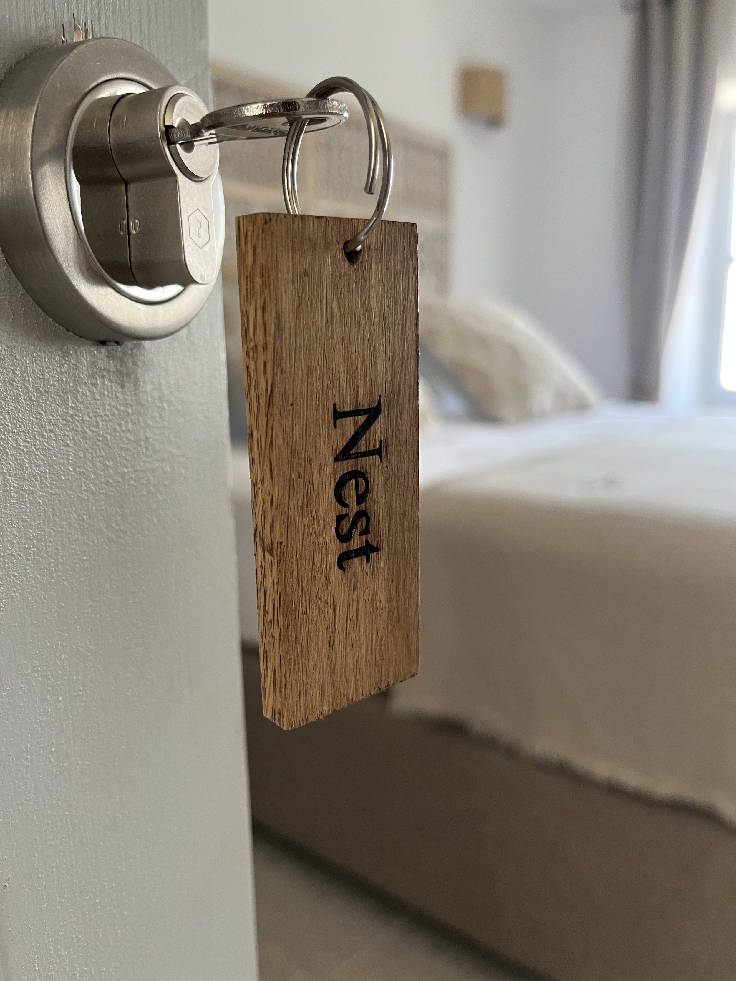 Close-up of a hotel room key card hanging on a silver keyring attached to a wall-mounted lock, with a bed and part of a window with curtains in the background.