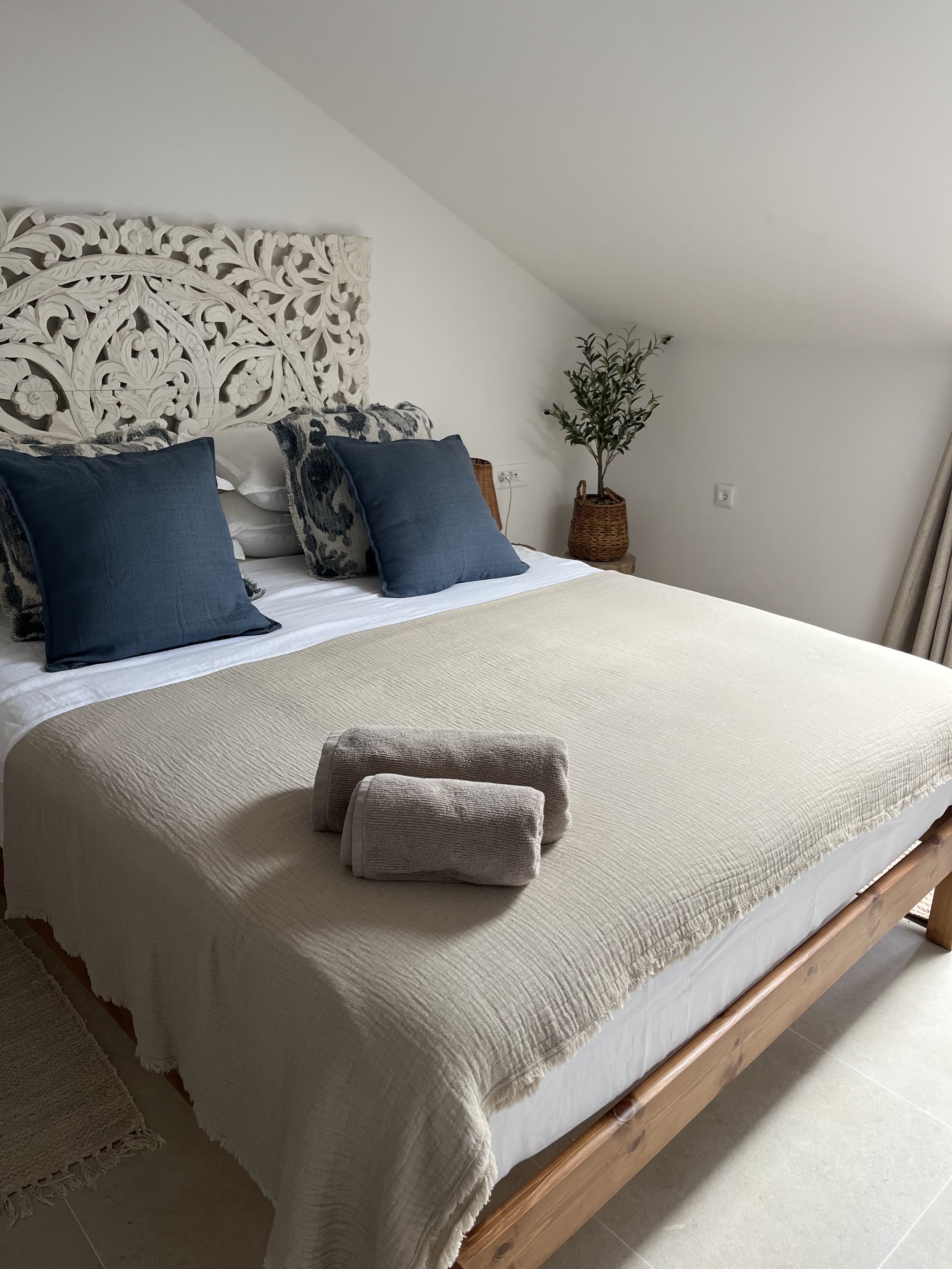 A neatly made bed with a beige quilt, dark blue pillows, and rolled towels on top, in a minimalist bedroom with a carved white headboard and a potted plant near the wall.