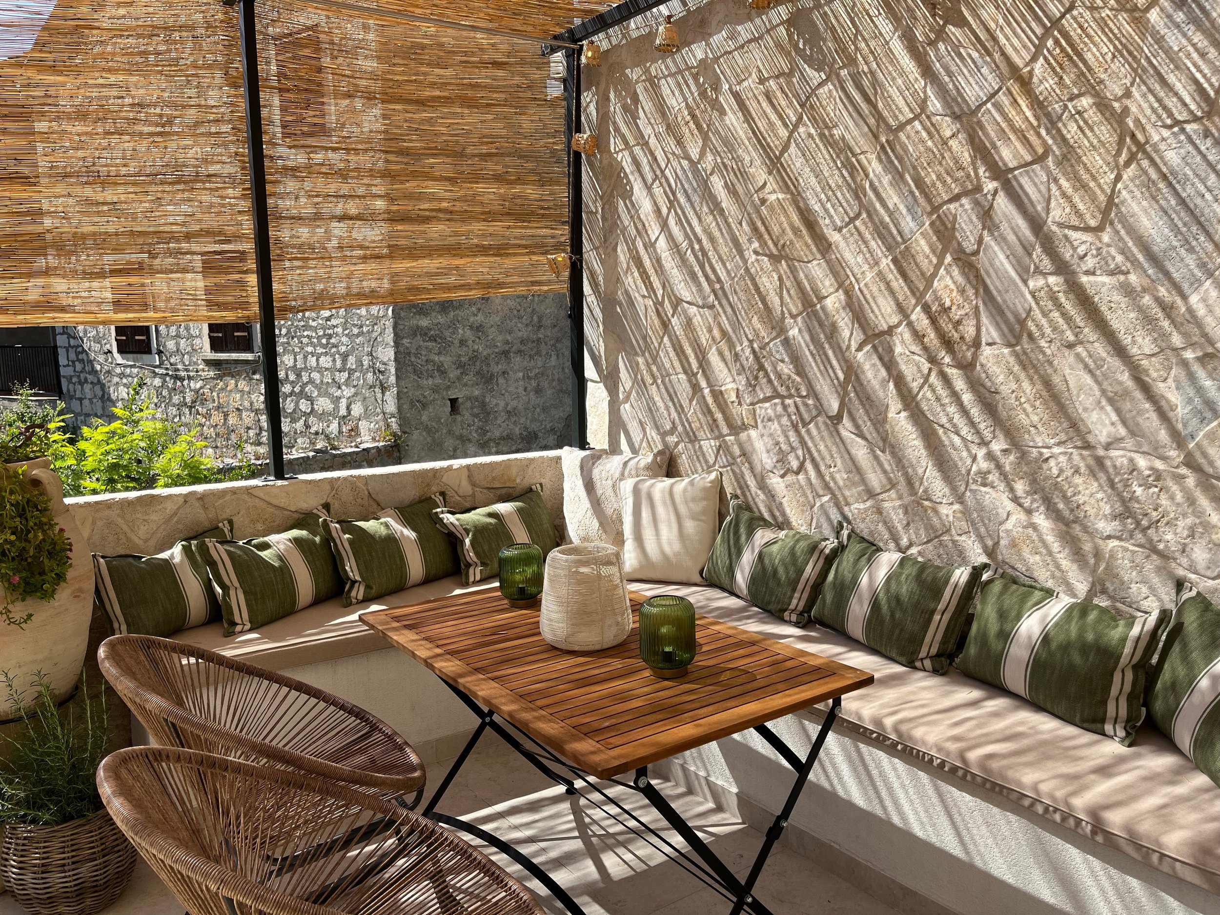 Outdoor patio with built-in cushioned seating along a stone wall, green striped pillows, a wooden table with decorative candles, wicker chairs, and a bamboo privacy screen with sunlight casting shadows.