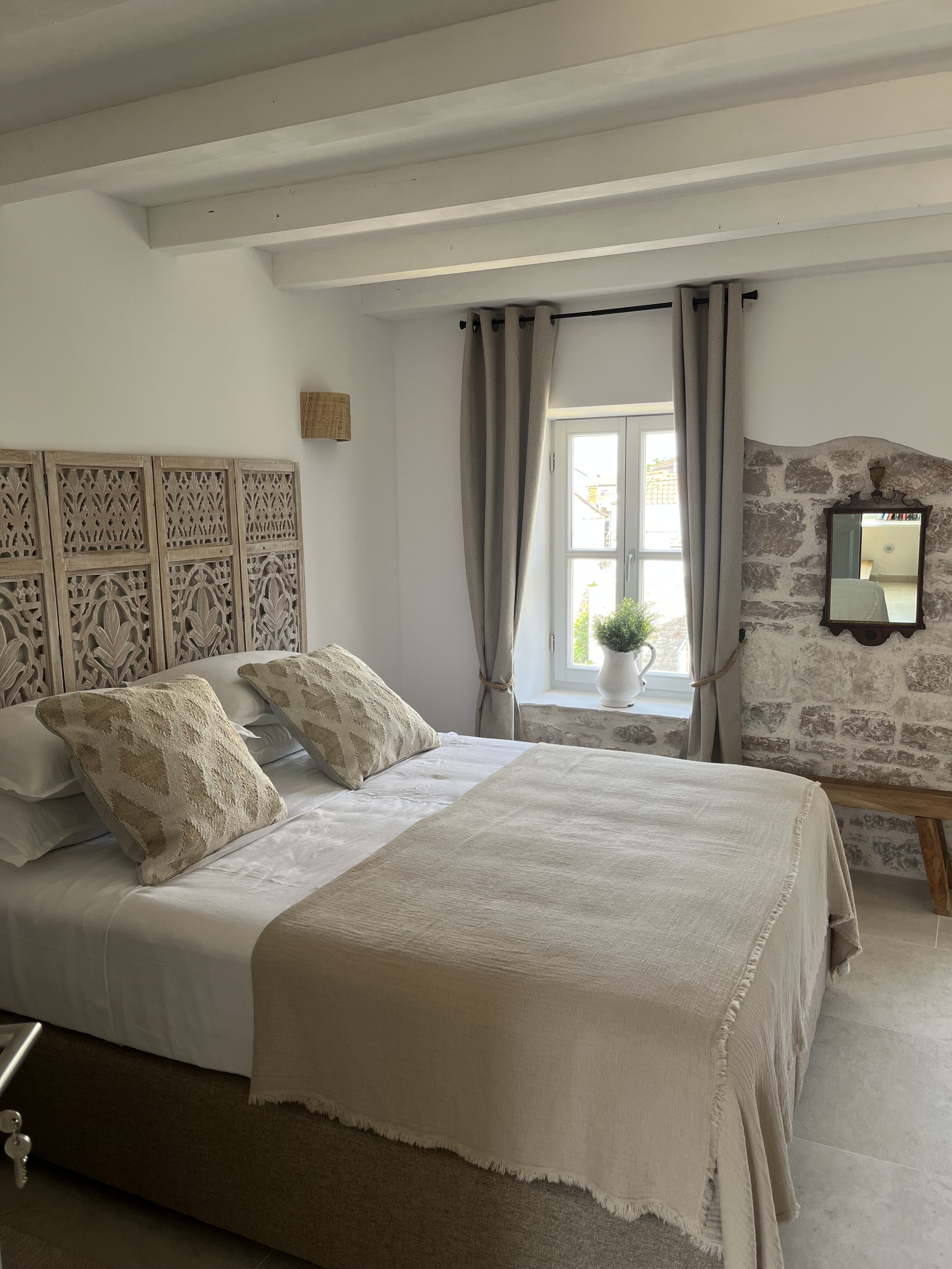 A cozy bedroom with a white bed, patterned pillows, a wooden headboard, a window with beige curtains, a small mirror on the wall, and a potted plant on the windowsill.