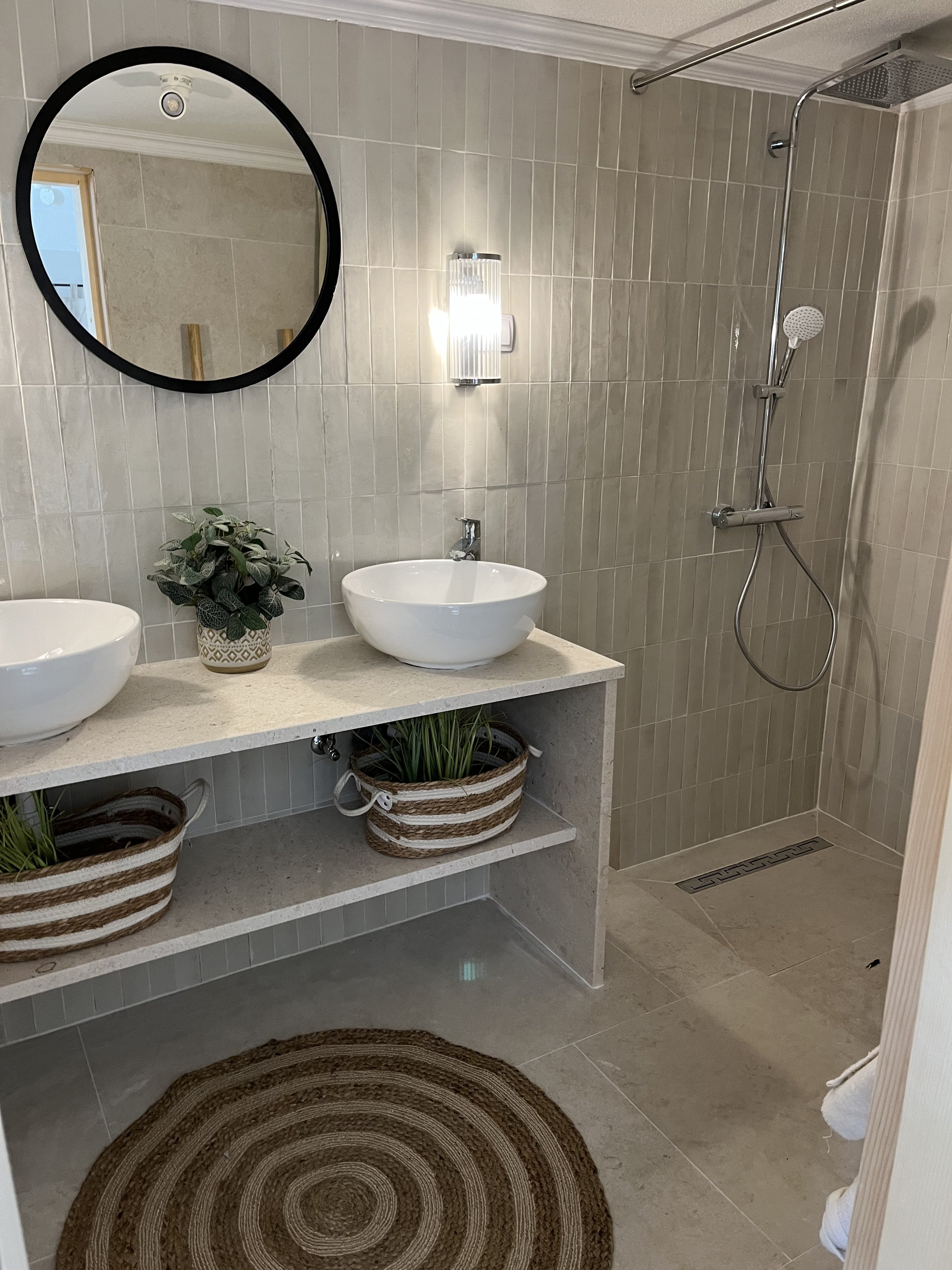 Bathroom with double vessel sinks on a light-colored stone countertop, a round mirror above, two woven baskets with plants underneath, a round rug on beige tile floor, beige tiled walk-in shower with handheld showerhead.
