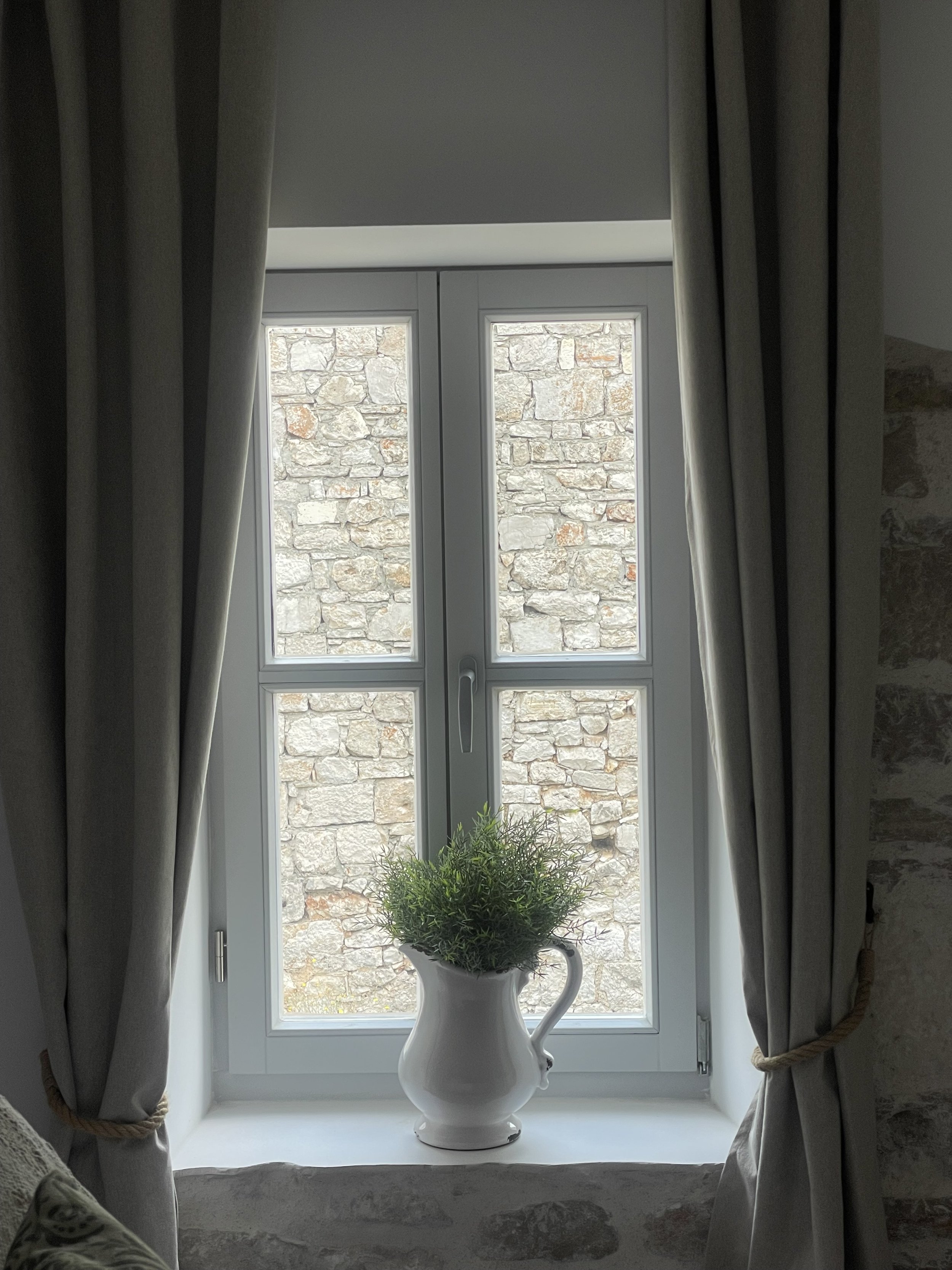 A window with stone wall outside, beige curtains tied back, and a white ceramic pitcher with greenery on the windowsill.