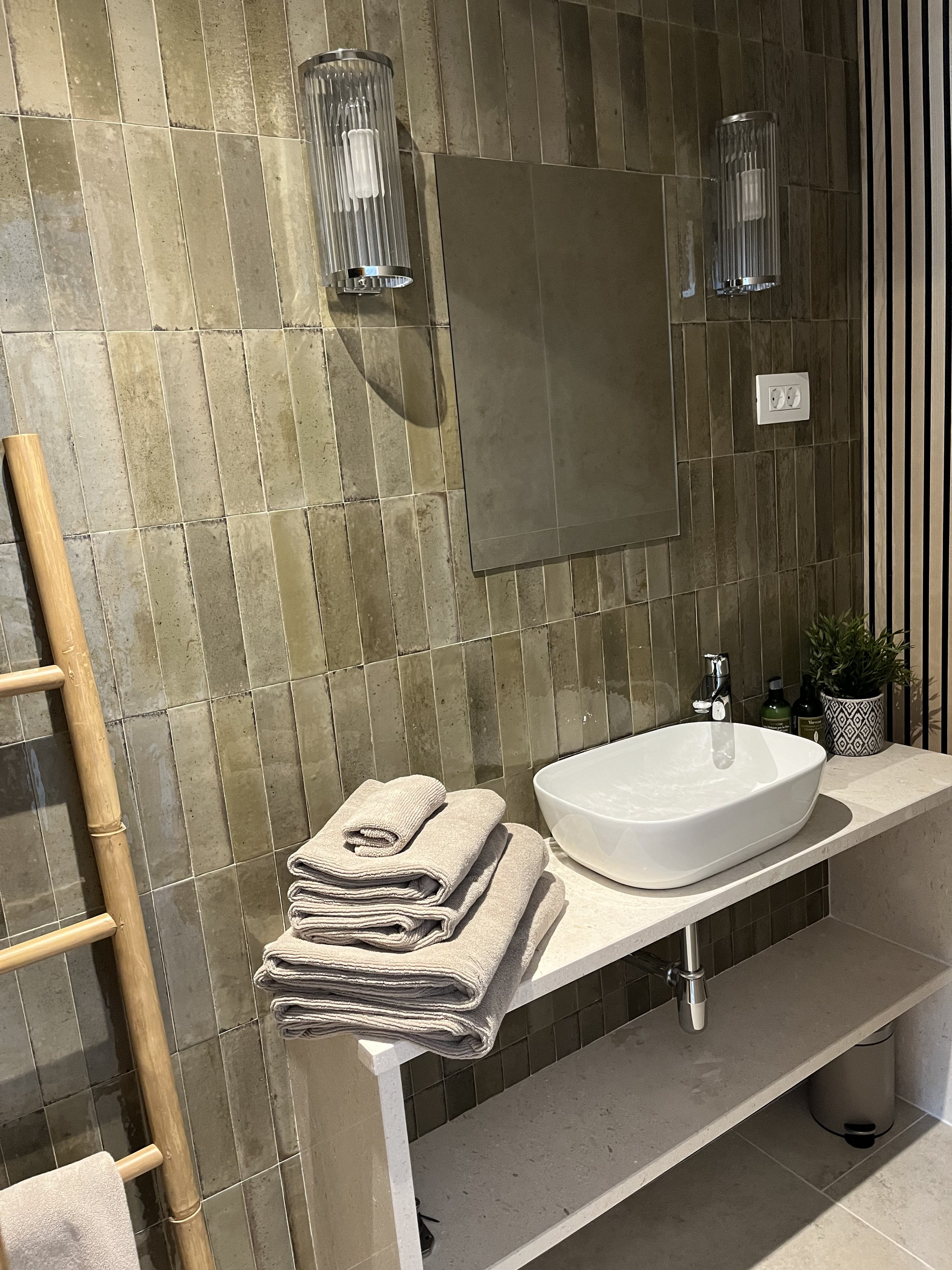 Bathroom with a white sink, stacked beige towels, green plants, and wall-mounted lights.