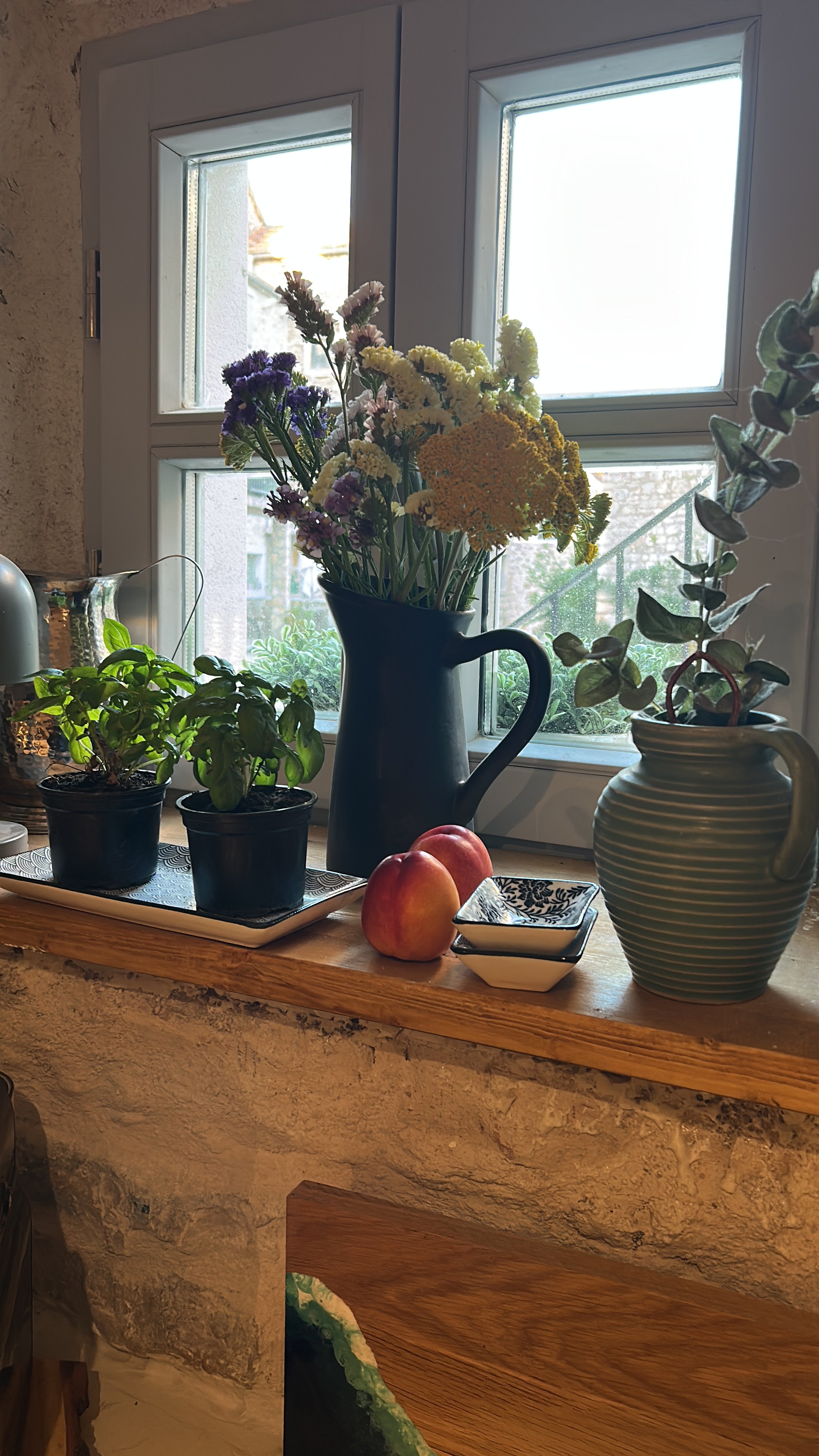 A kitchen windowsill with potted herbs, a black vase with a colorful flower bouquet, a striped green vase with an eucalyptus branch, two apples, and a small square dish.