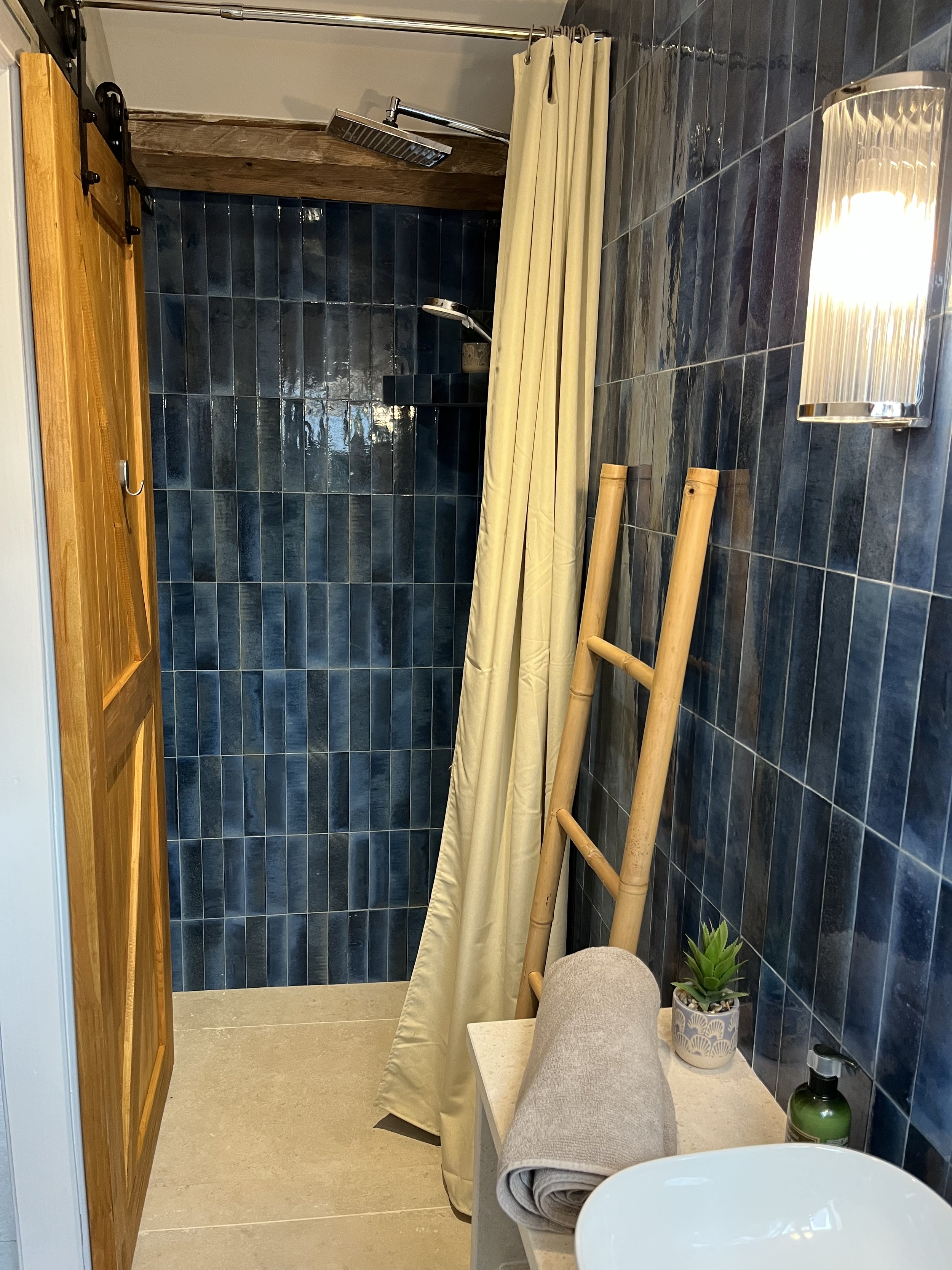Bathroom with a walk-in shower covered by a yellow shower curtain, dark blue tiled walls, wooden accents, a ladder, a potted plant on a small table, a rolled towel, and a soap dispenser.