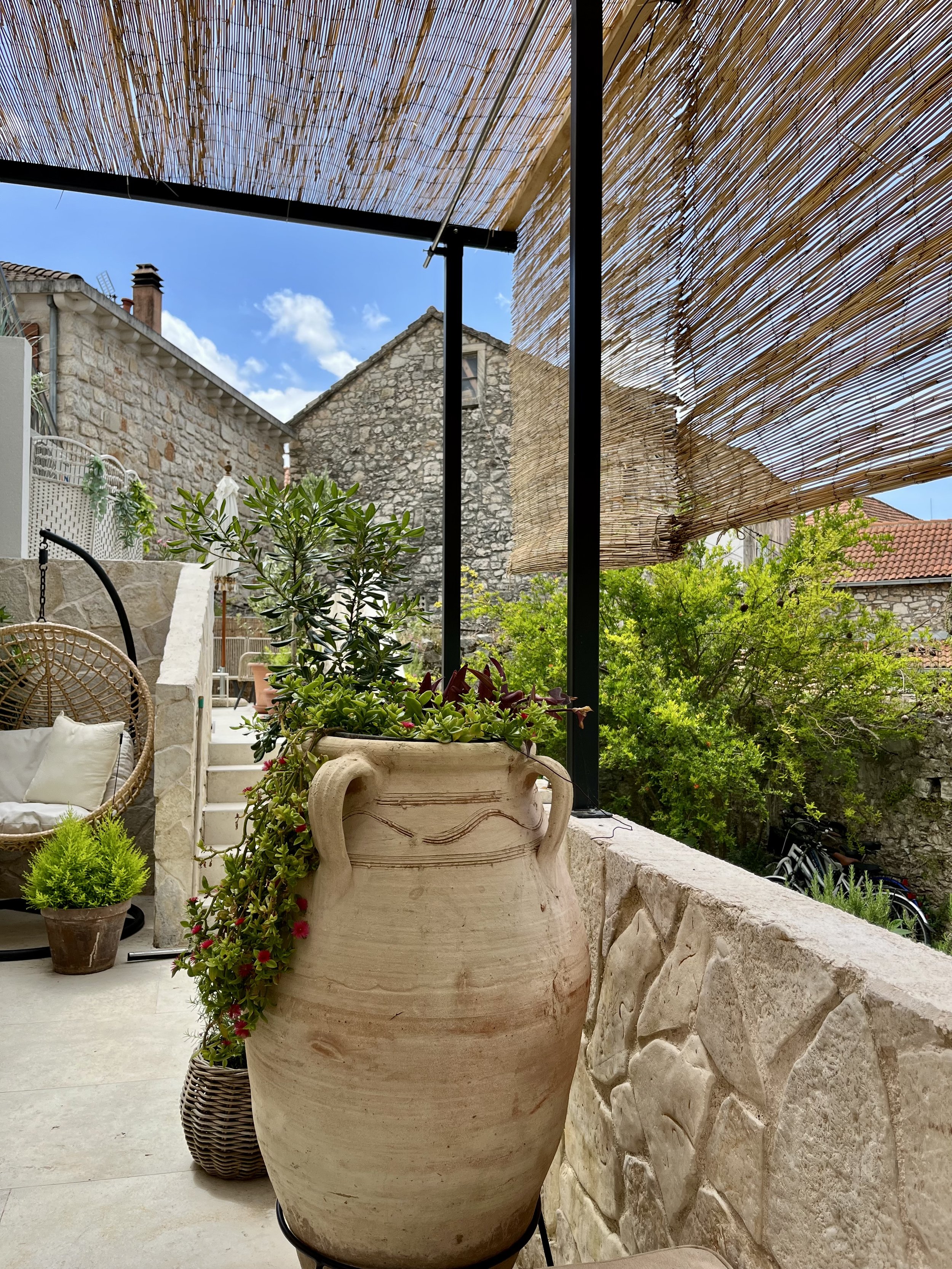 Outdoor patio with large ceramic pot filled with green plants, stone wall, wicker swing with cushions, potted plant, lush greenery, and a wooden sunshade under a blue sky with clouds.