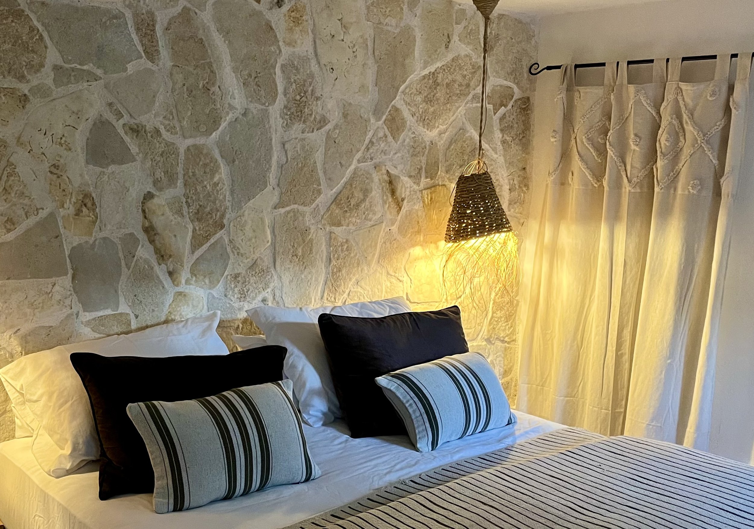 A bedroom with a stone accent wall, a bed with white sheets and multiple pillows, and a window with cream-colored curtains. A woven hanging lamp emits warm light beside the bed.