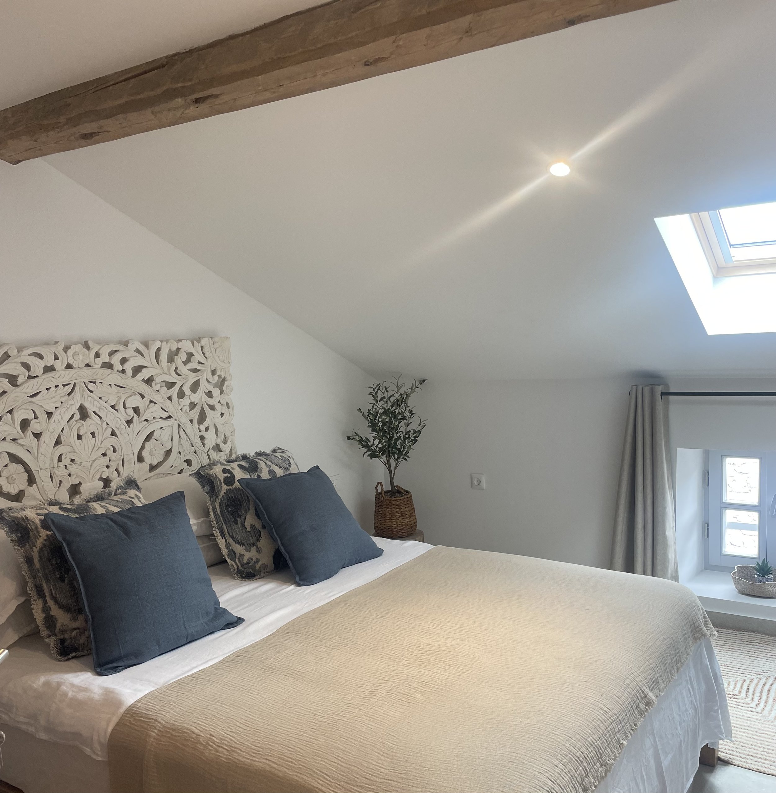 Cozy bedroom with a large bed featuring a carved white headboard, navy blue and patterned pillows, a beige bedspread, potted plant on a nightstand, skylight, small window with curtains, and a sloped ceiling with a wooden beam.