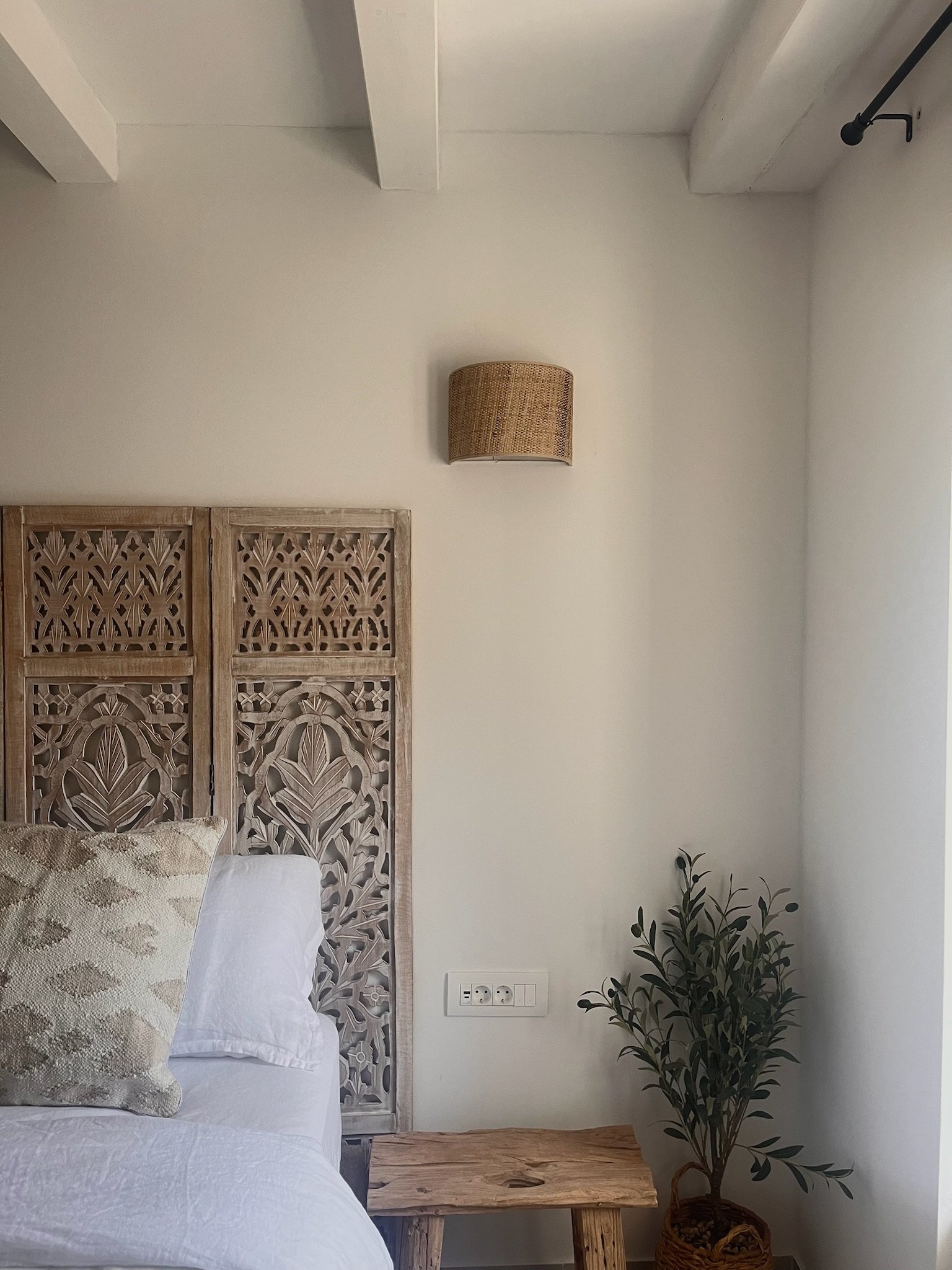 A bedroom corner with a wooden headboard, a white bed with a beige pillow, a wooden side table, a potted plant, a wall-mounted light fixture, and a window with a curtain rod.