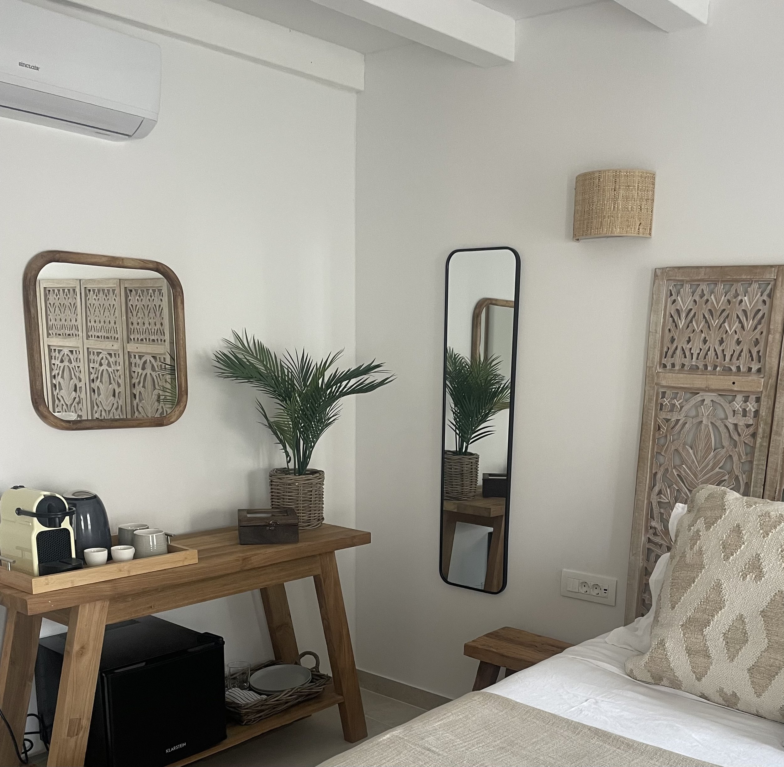 A cozy hotel room corner with a wooden table holding a coffee machine, cups, a potted plant, and a tissue box. A mirror, wall-mounted light, decorative wooden panel, and bed with patterned pillows are also visible.