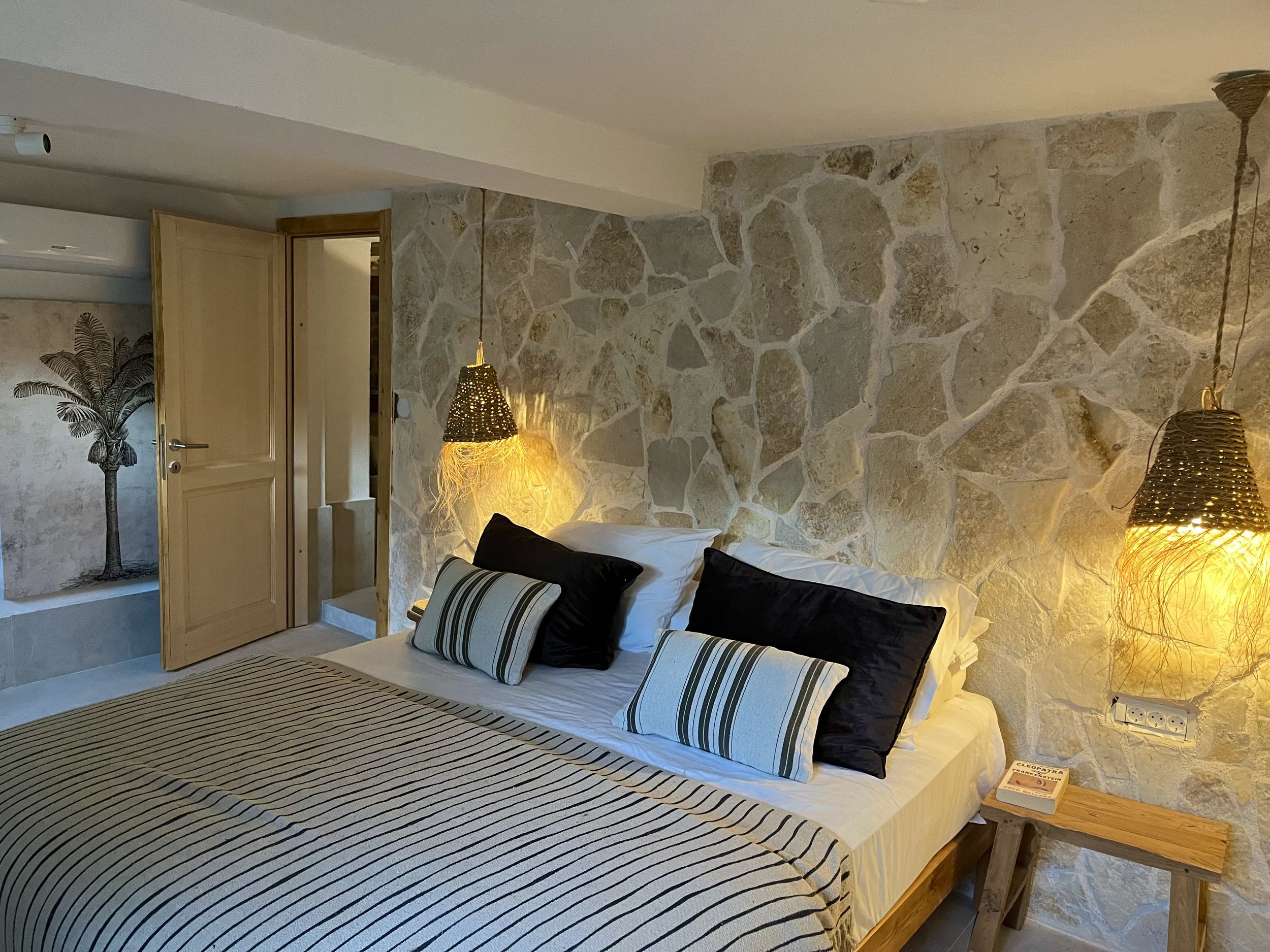 A bedroom with a stone wall, bed with white and black pillows, striped bedspread, wooden nightstand, and hanging wicker lamps.