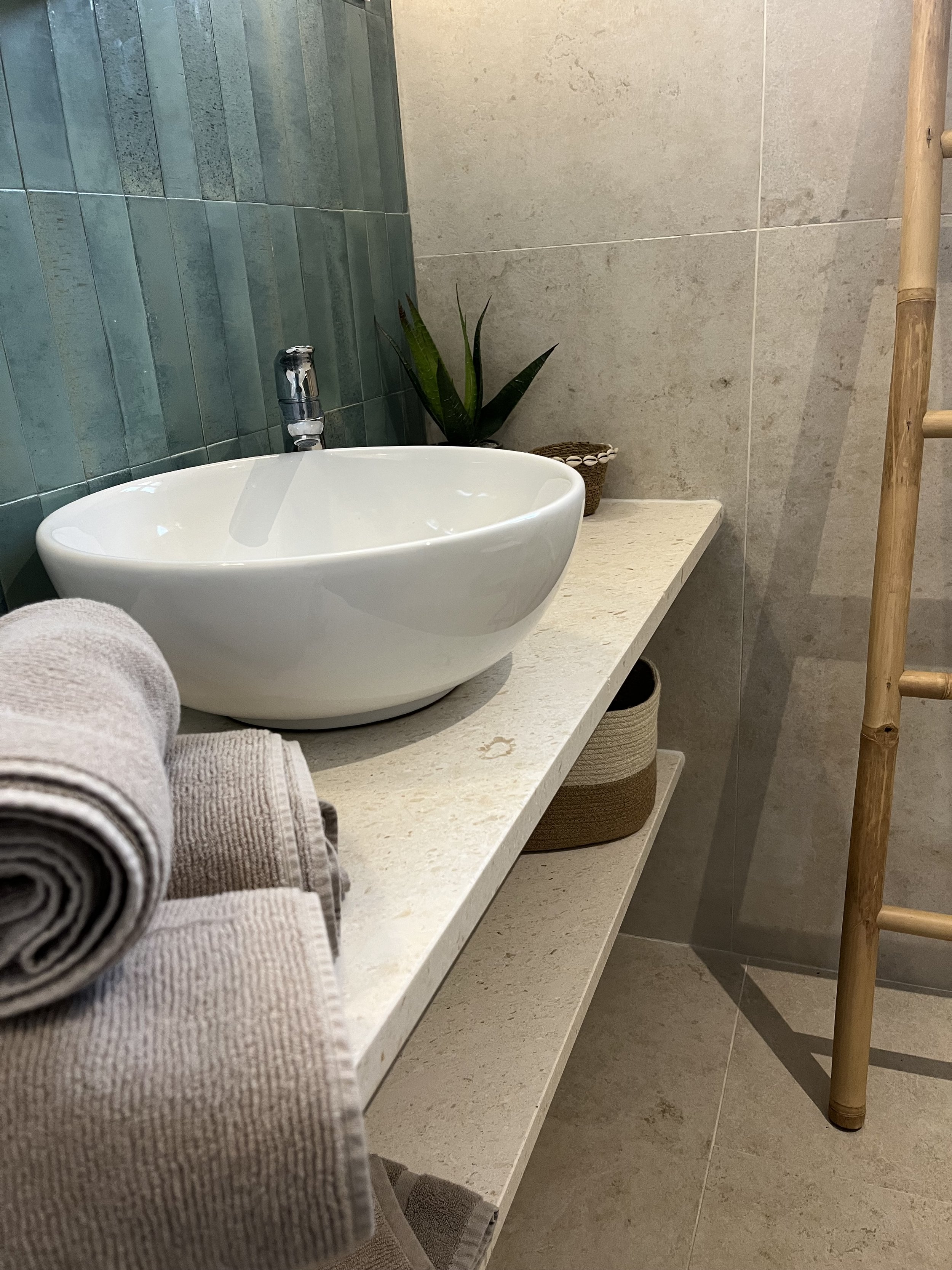A bathroom countertop with a white oval sink, a modern chrome faucet, and a small potted plant. There are neatly rolled towels on the left, and a basket with a plant behind the sink. A bamboo ladder is on the right.
