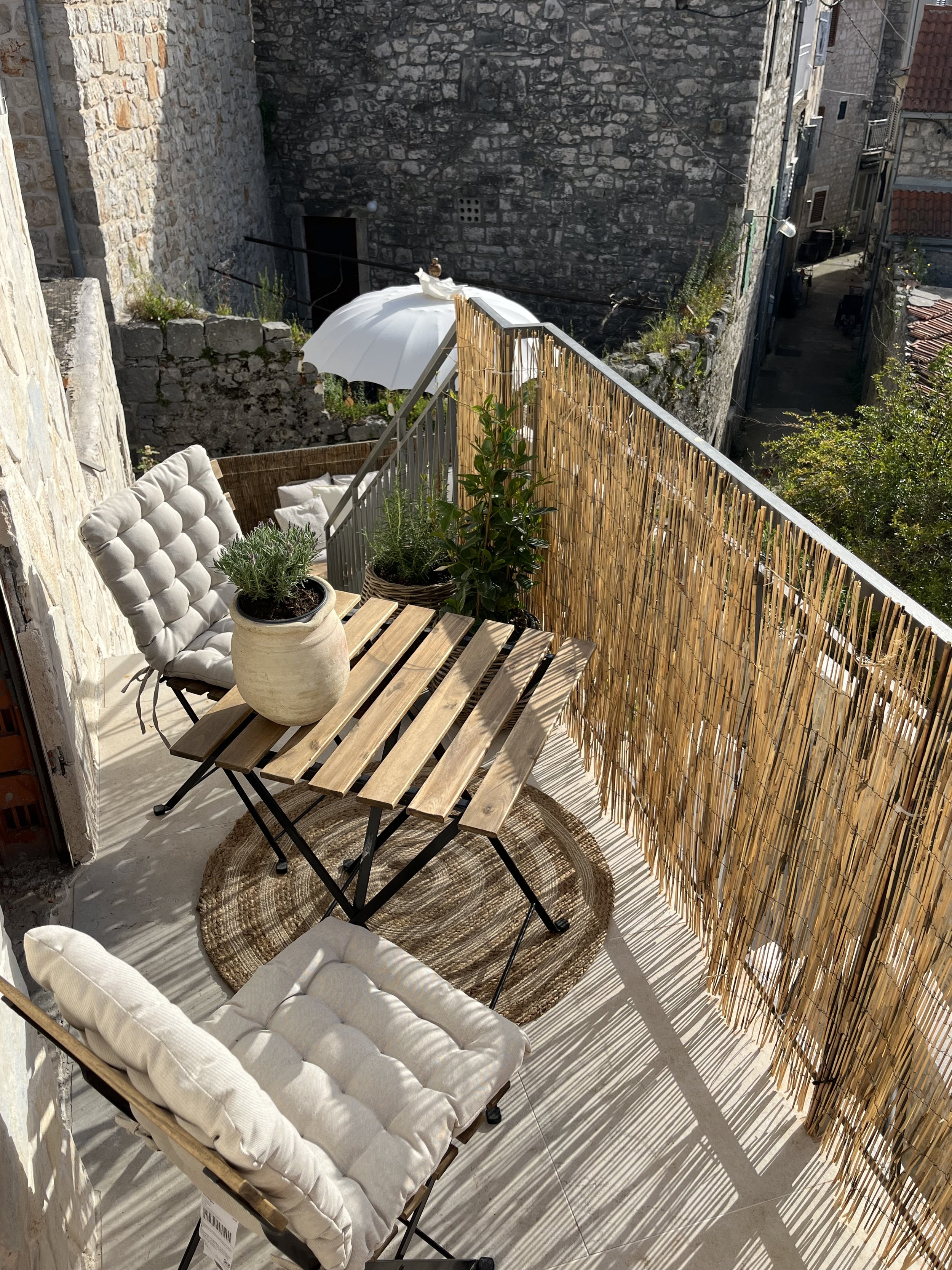 Small balcony with wooden table and two cushioned chairs, potted plants, a white umbrella, and bamboo privacy screen, overlooking a narrow street with stone buildings.
