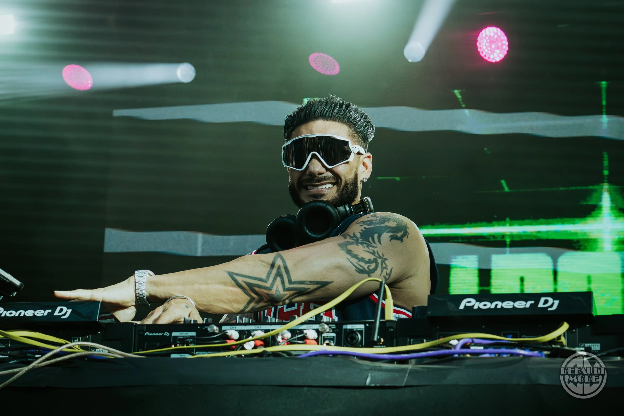 A smiling male DJ wearing sunglasses and headphones around his neck, standing behind a Pioneer DJ setup with wires, in a club with colorful lights and a digital screen in the background.