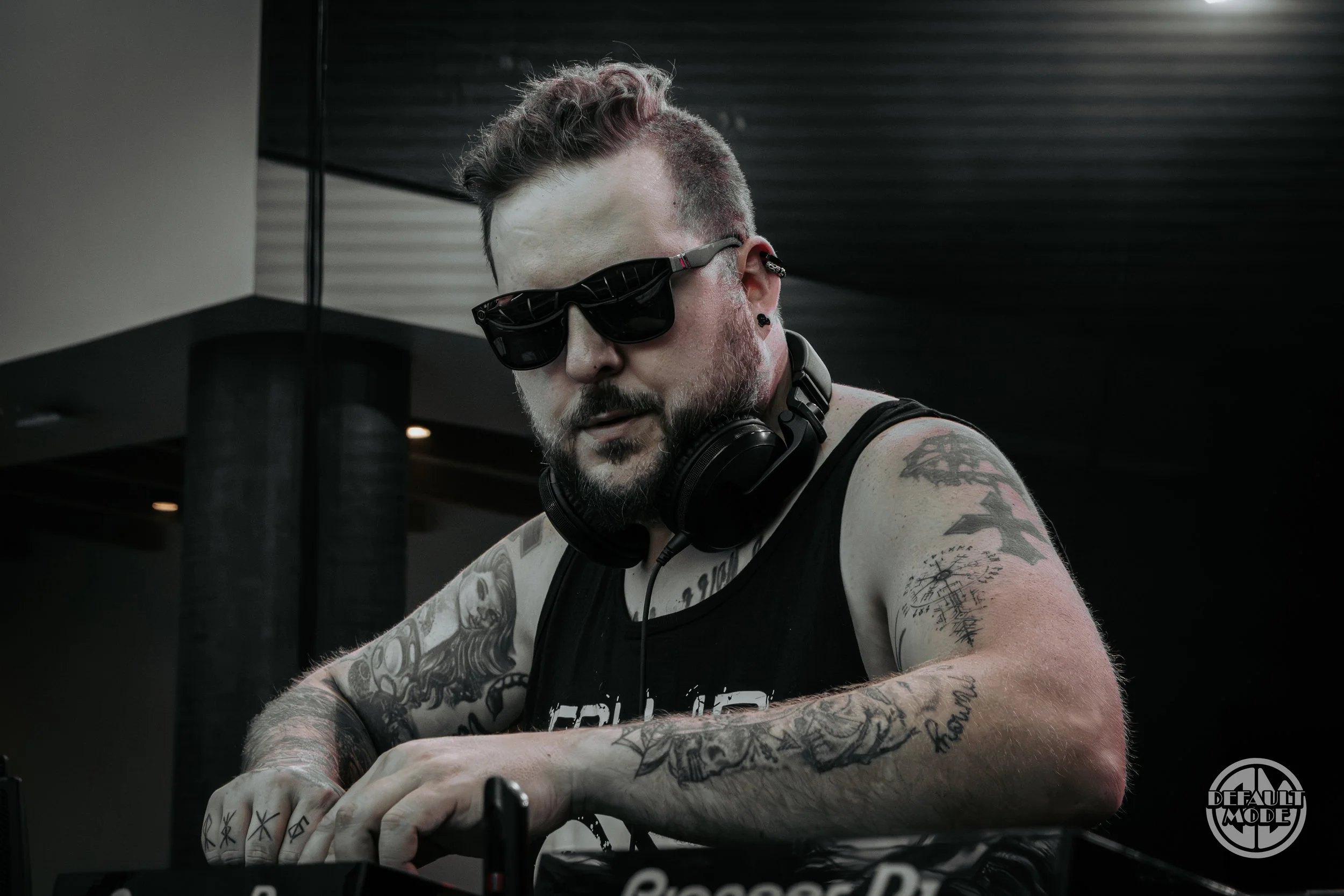 A tattooed DJ wearing sunglasses and headphones around his neck working with DJ equipment in a dimly lit space.