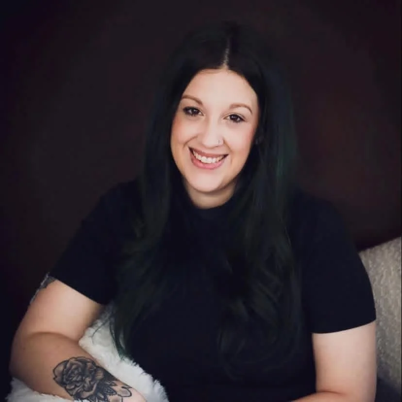 A woman with green hair, smiling widely, is holding a small brown and black dog. The woman is wearing a black sheer top with spiderweb patterns and a black skirt. They are indoors, with a plain wall and wooden floor in the background.